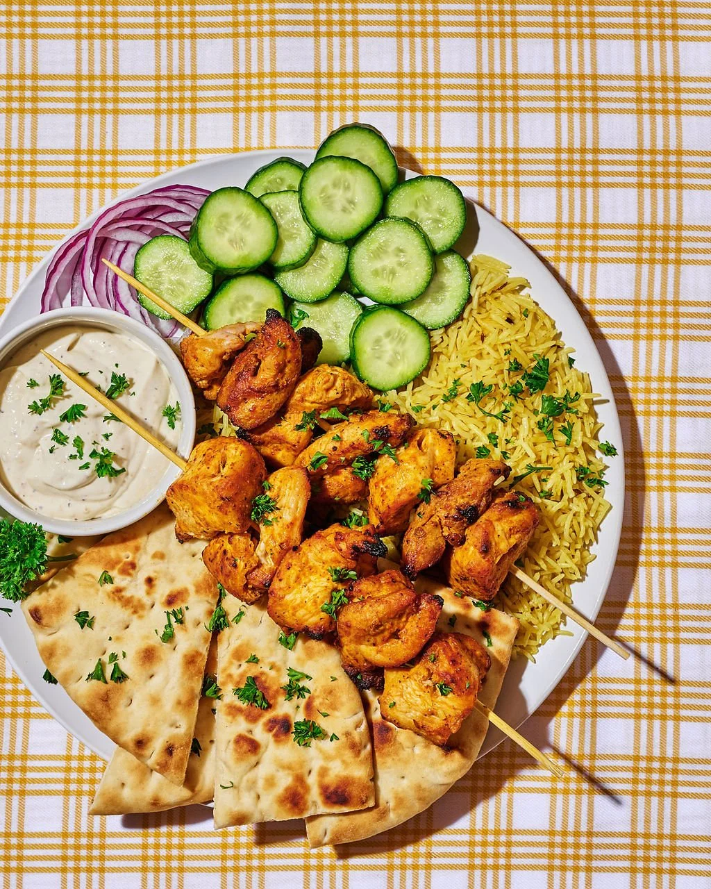 Plate of chicken kebabs with naan, yellow rice, sliced cucumbers, red onion, and a side of creamy dipping sauce garnished with parsley, on a yellow checkered tablecloth.