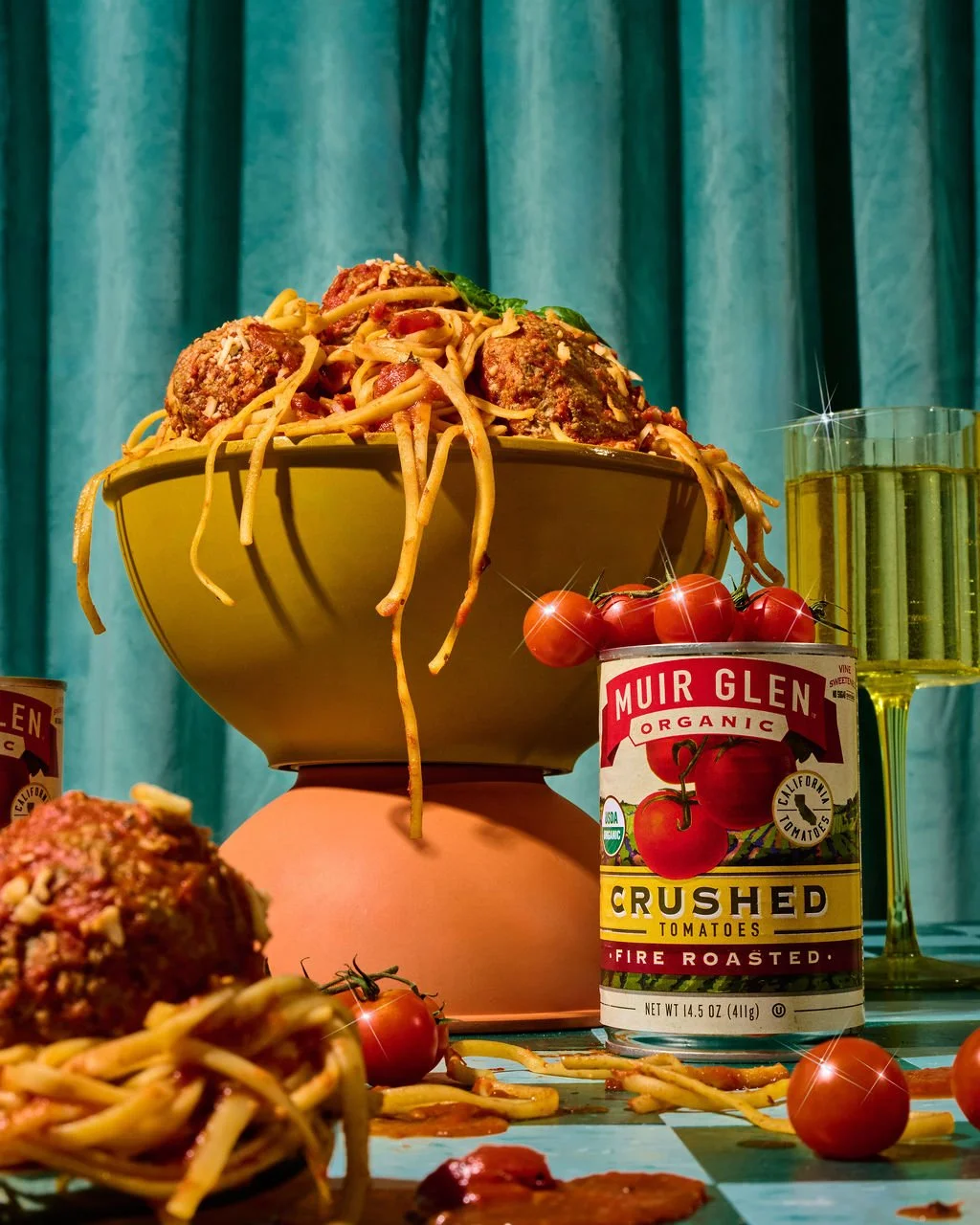 A bowl of spaghetti with meatballs and tomato sauce on a pink stand, with cherry tomatoes, a can of crushed tomatoes, and a glass of white wine on a table with a blue curtain background.
