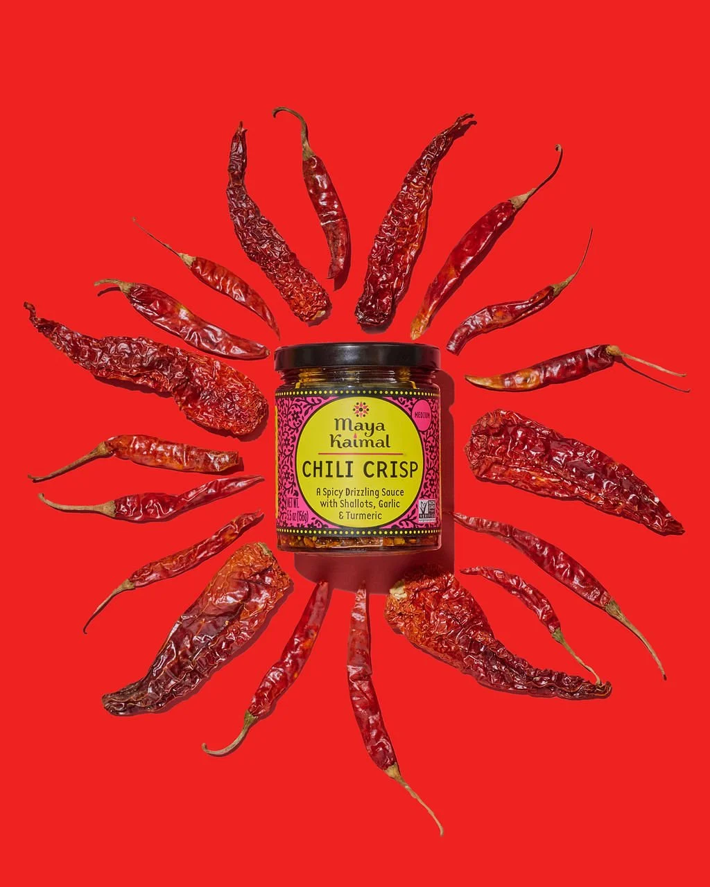 A jar of Maya Kaimal Chili Crisp sauce surrounded by dried red chili peppers on a red background.