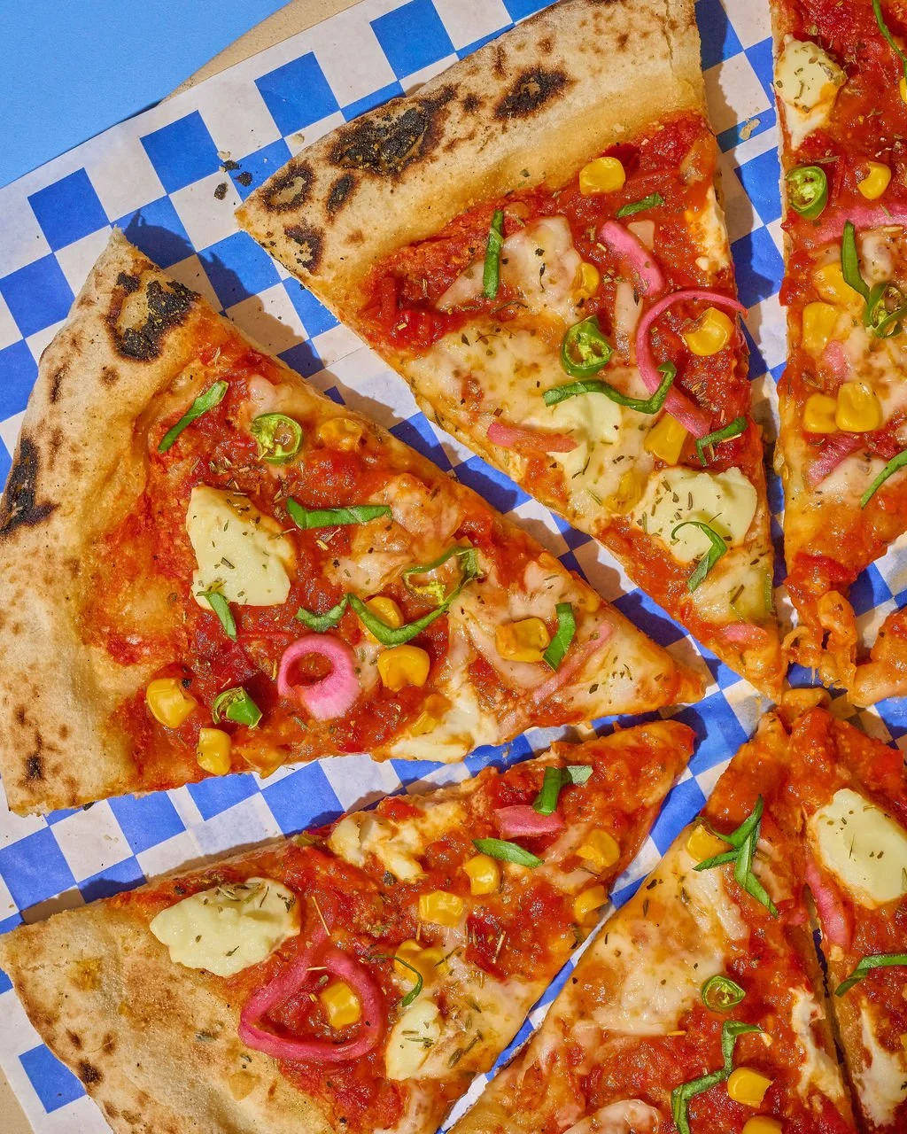 Close-up of multiple slices of vegetable pizza with toppings like green peppers, corn, pickles, and pink onions on a checkered blue and white paper.