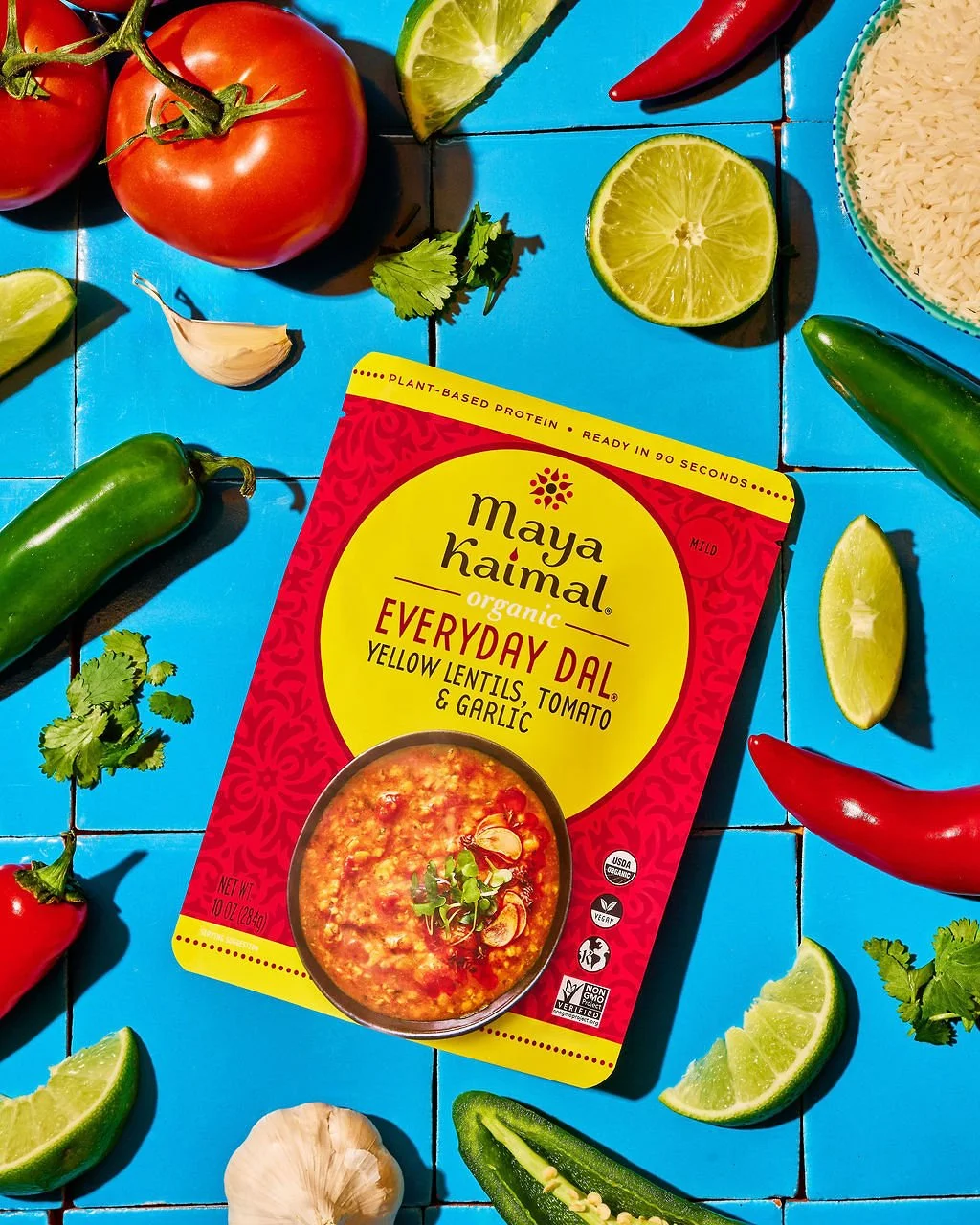 Maya Kaimal organic everyday dal box surrounded by fresh tomatoes, lime halves, a garlic clove, jalapeno pepper, cilantro, and rice on a blue tiled surface.