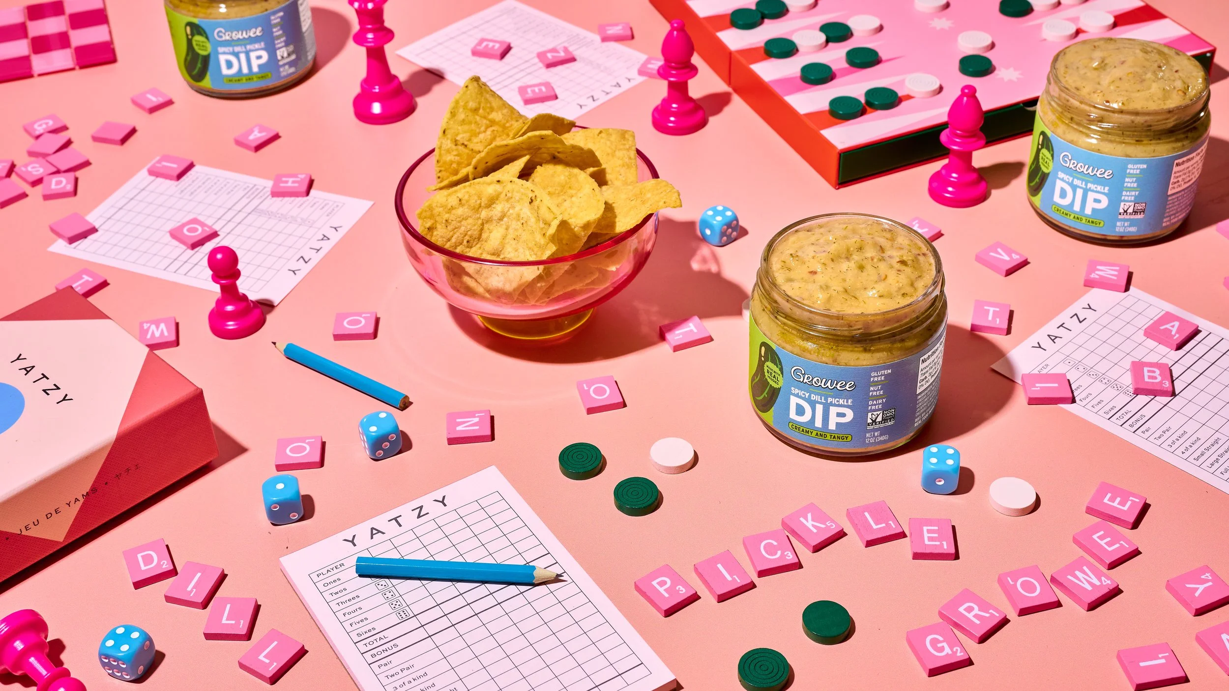 Table set for a game night with various board game pieces, Scrabble tiles, and bowls of spicy dill pickle dip on a pink table.