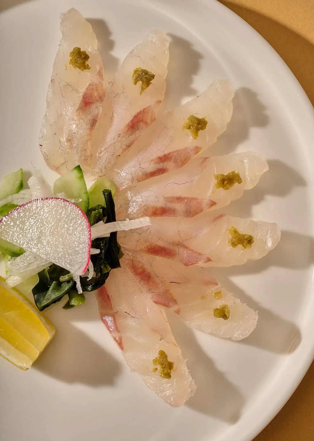 Thin slices of sashimi (raw fish) with a small amount of wasabi on top, garnished with vegetables, lemon, and radish on a white plate.