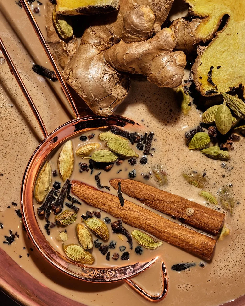 Close-up of a spice mixture in a copper spoon, containing cardamom pods, cinnamon sticks, black peppercorns, and cloves, surrounded by chunks of ginger and turmeric root in a creamy liquid.