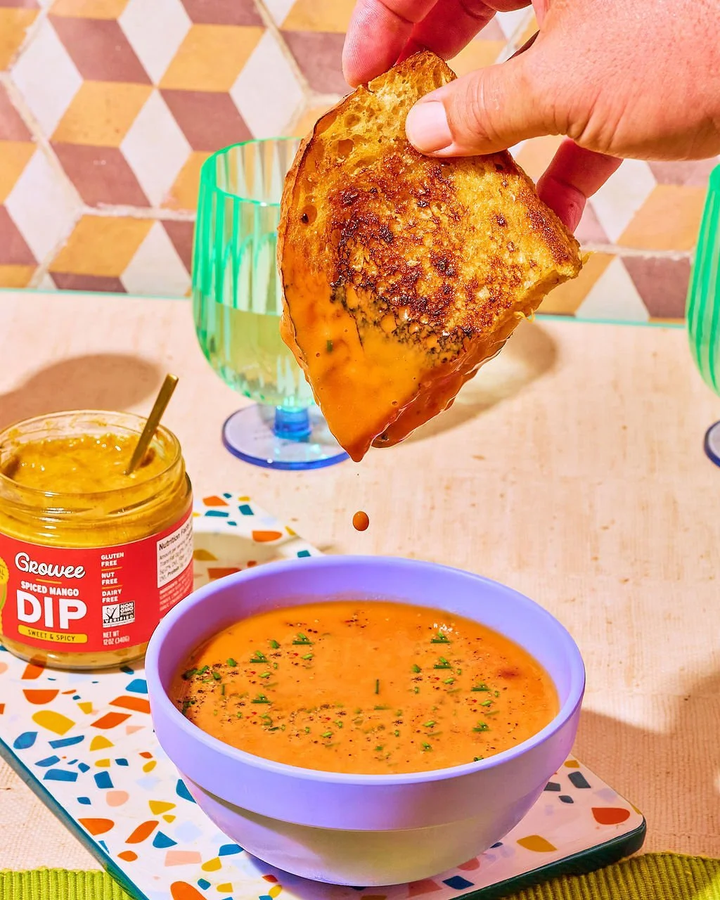 A hand holds a piece of toasted bread over a bowl of soup, with a jar of mango dip and two glasses in the background.