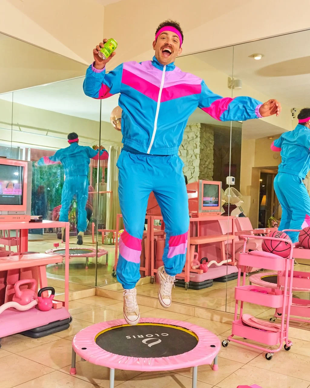 Man in colorful 80s workout attire jumping on a mini trampoline in a room decorated with pink fitness equipment and retro electronics.