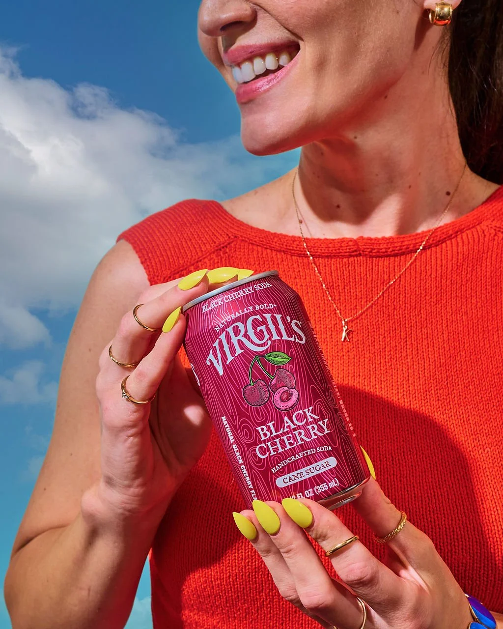 A woman in a red sleeveless top holding a can of Virgils Black Cherry handcrafted soda, with a bright smile, against a background of blue sky and clouds.