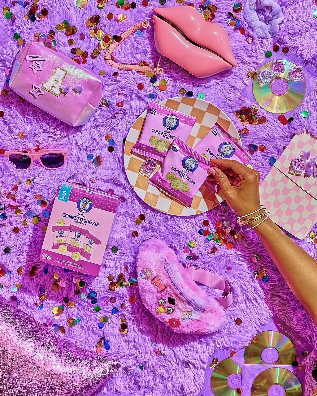 Pink and purple party items including confetti, a pink lips-shaped purse, sunglasses, fuzzy stuffed toy, mini confetti sugar packets, CDs, a pink pouch, and a purple checkered box, all on a purple shaggy surface with scattered colorful confetti.