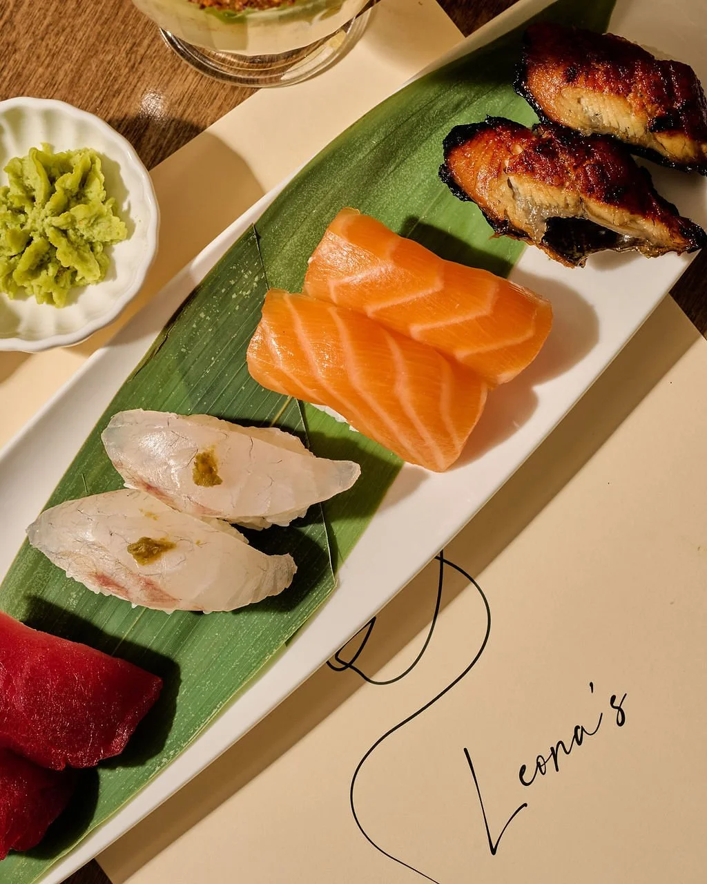 Assorted sashimi including salmon, tuna, and white fish, with wasabi on the side, served on a banana leaf on a white platter.