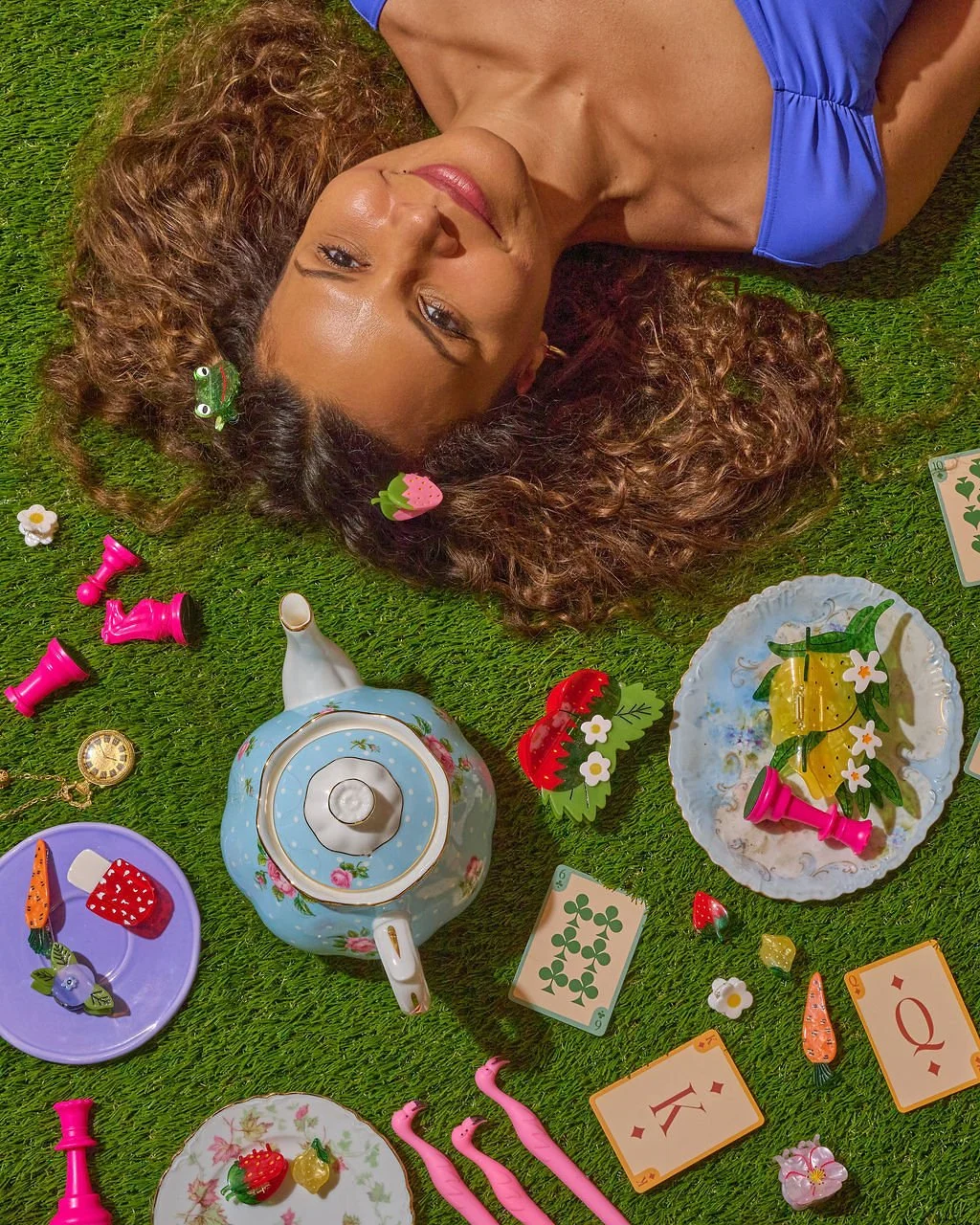 A woman with curly hair lying on green grass, wearing a blue top, surrounded by a teapot, plates with desserts, cards, and small decorative items.