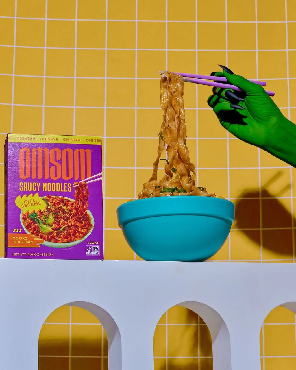 A person with a green glove holding purple chopsticks lifts cooked noodles from a blue bowl, with a box of OMISOM saucy noodles on a yellow tiled wall background.