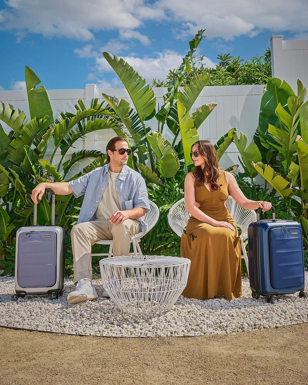 A man and woman sitting outdoors on white chairs with suitcases beside them, surrounded by tropical plants and a white fence under a blue sky.