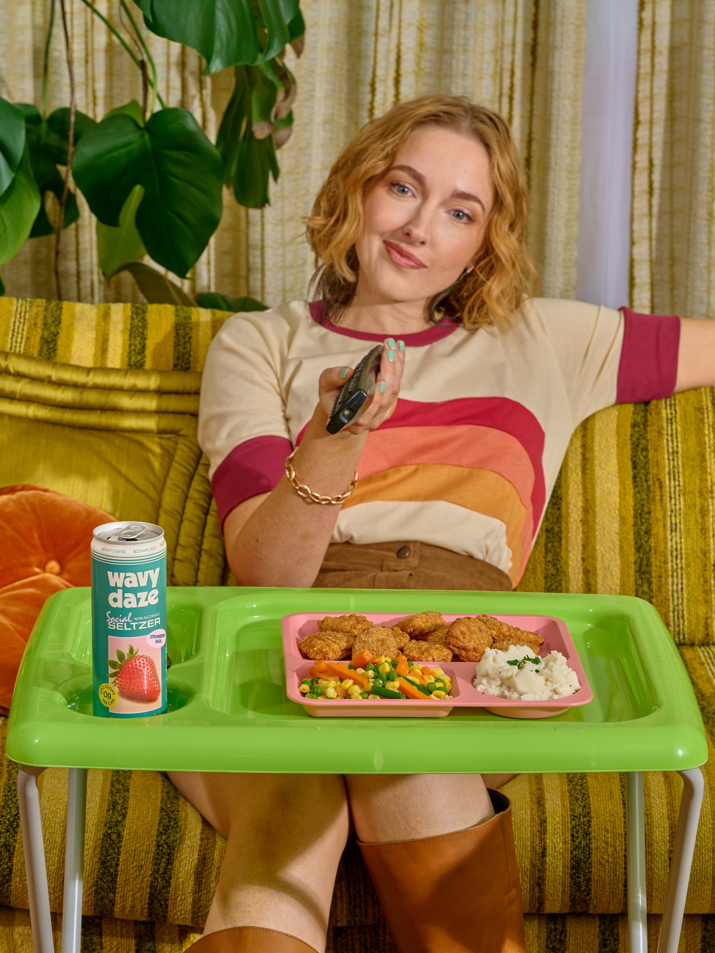 A woman sitting on a vintage yellow-striped sofa, holding a TV remote, with a green snack tray on her lap containing chicken nuggets, mixed vegetables, and mashed potatoes. There is a can of strawberry flavored seltzer on the tray and large green plants in the background.