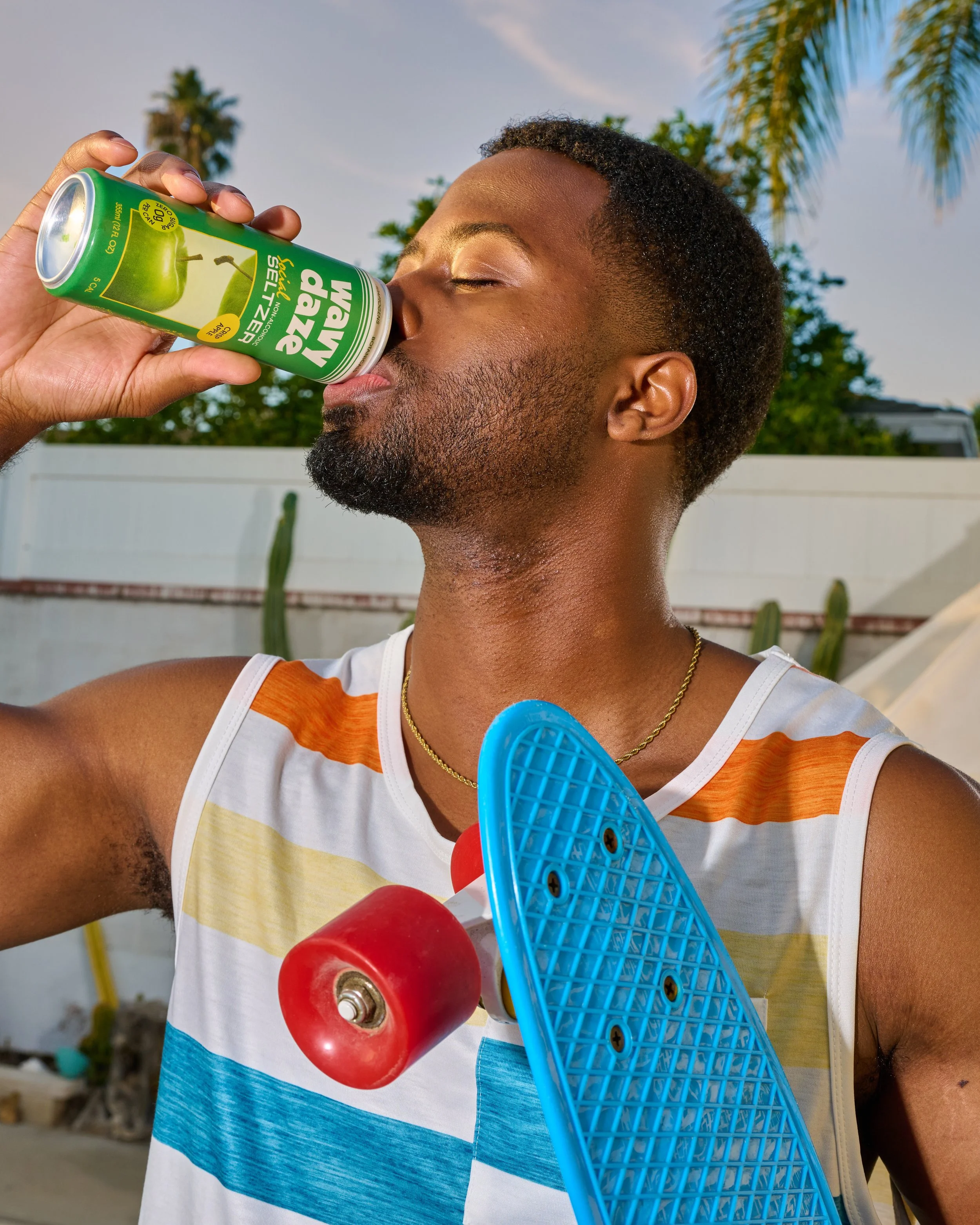 A young man in a colorful striped tank top drinking watermelon flavored sparkling water from a can outdoors, with a skateboard hanging from his shoulder and palm trees in the background.