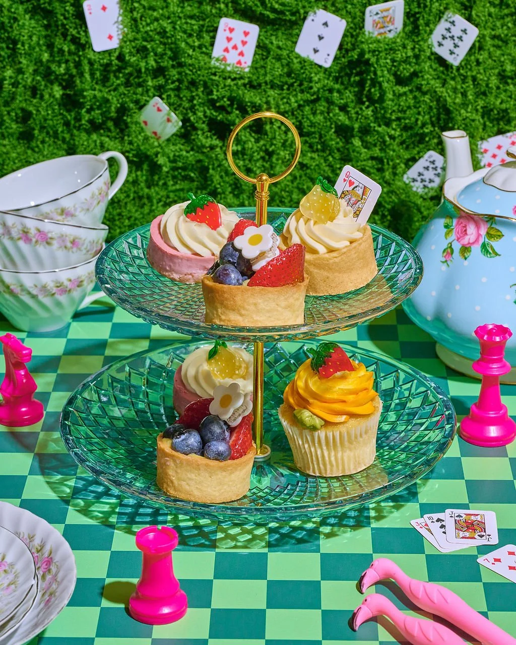A colorful tea party setup with two-tiered glass serving trays holding fruit-topped mini cheesecakes, surrounded by floral china teacups and a teapot, pink chess pieces, playing cards, and a green checkered tablecloth, set against a grassy background
