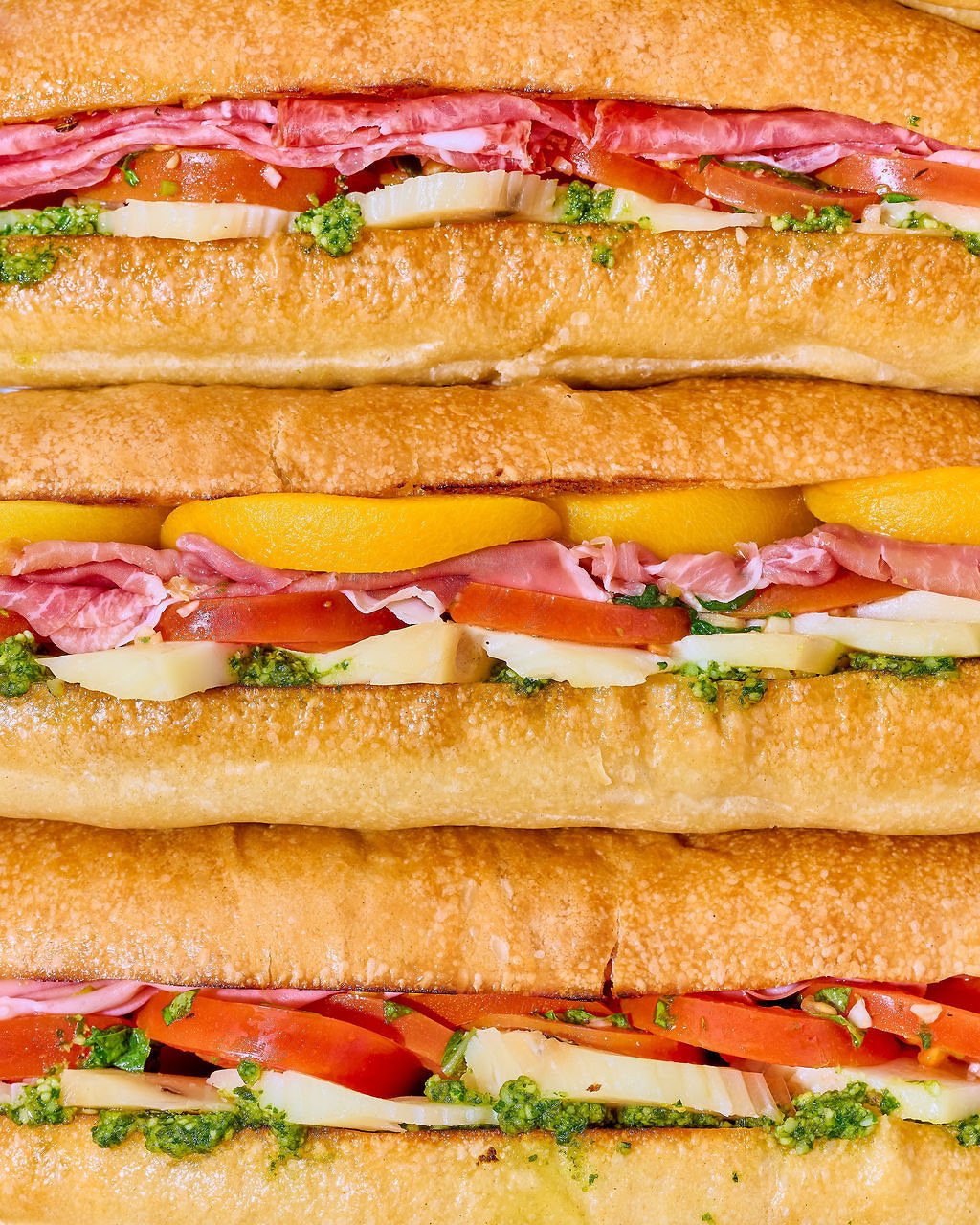 Close-up of multiple Italian hoagie sandwiches filled with ham, beef, tomatoes, cheese, lettuce, and green sauce, in toasted rolls.