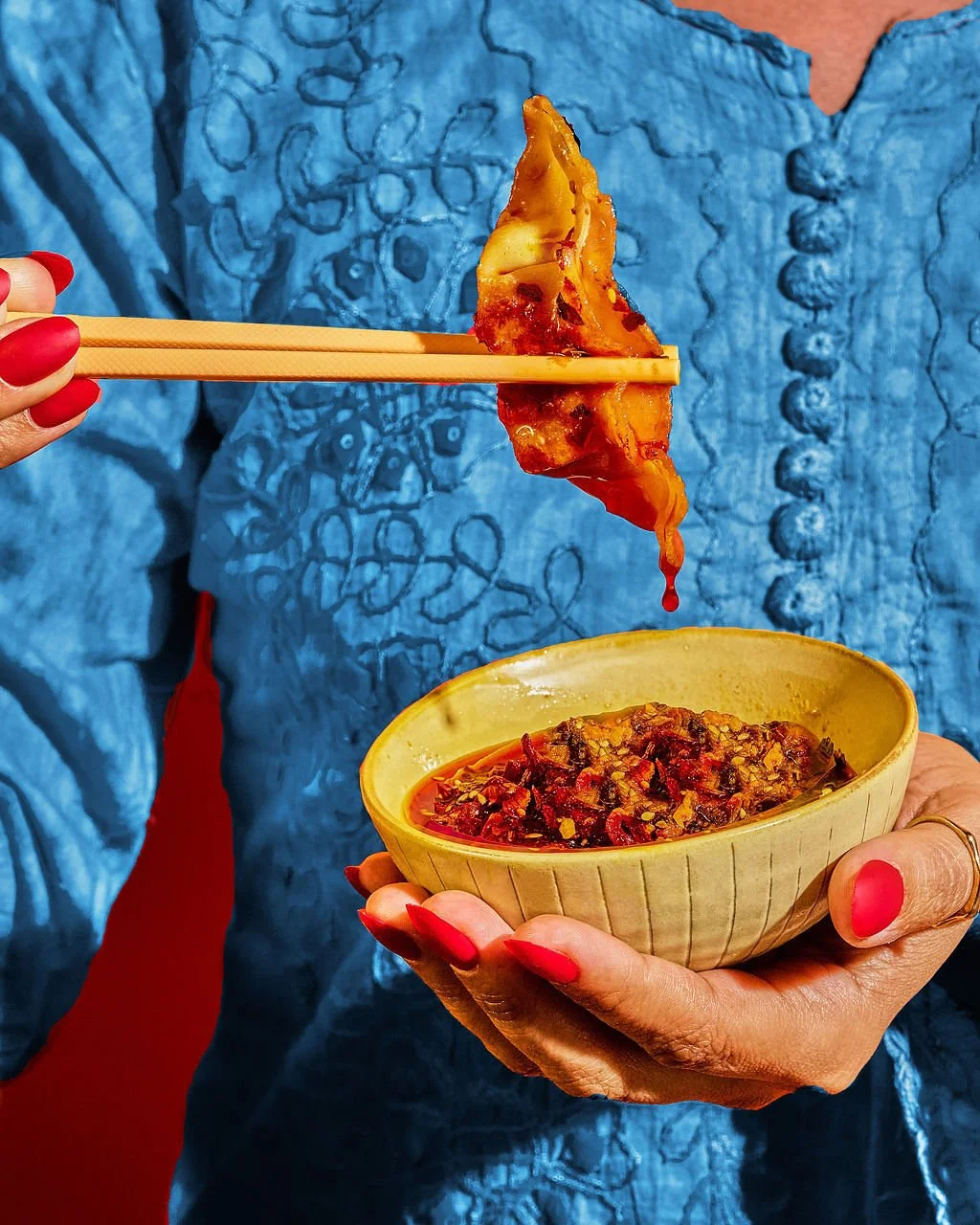 Person with red nail polish picking up a piece of food with chopsticks from a yellow bowl of chili or spicy stew.