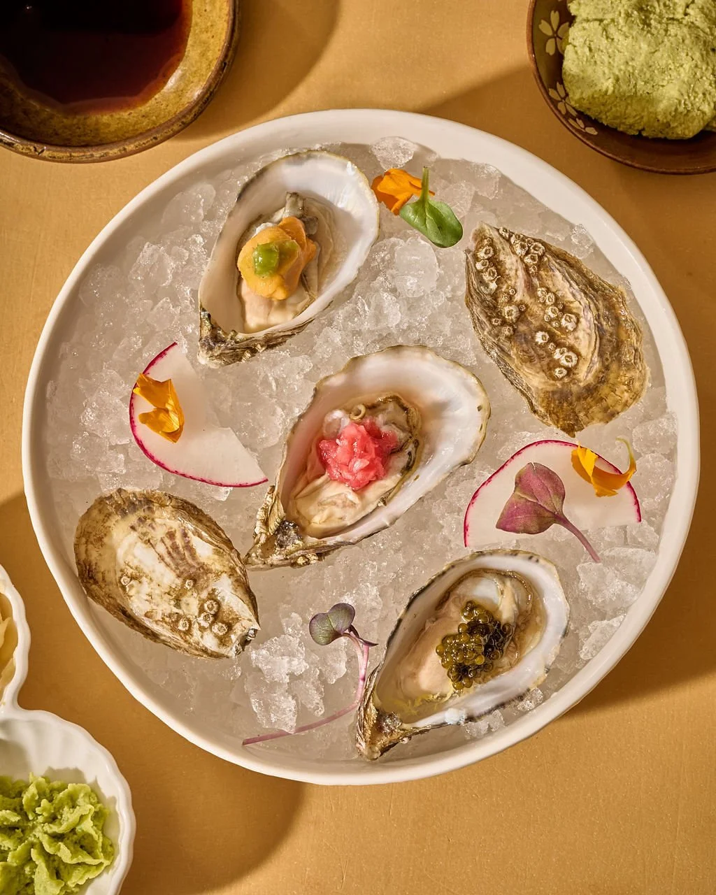 A white platter of fresh oysters on ice, garnished with edible flowers and small piles of condiments, with a bowl of wasabi and a drink in the background.