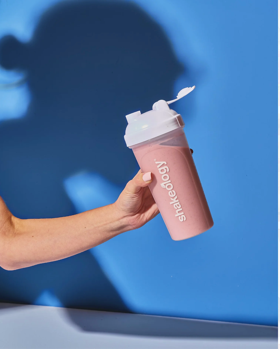 A person holding a pink Shakeology shaker cup with the logo visible, featuring a white lid open with a flip-top cap. The person's arm and hand are visible, with a light skin tone and painted nails. The background is blue with a clear shadow of the person's head and shoulder cast on it.
