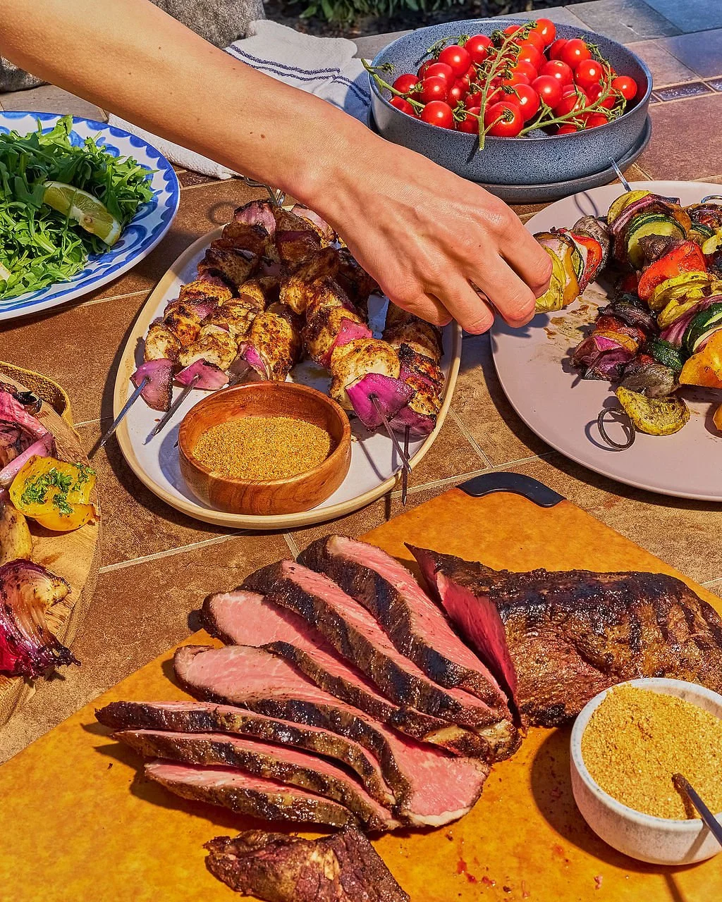 Sliced grilled steak, skewers of cooked meat and vegetables, cherry tomatoes, salad, and slices of grilled meat on a table.