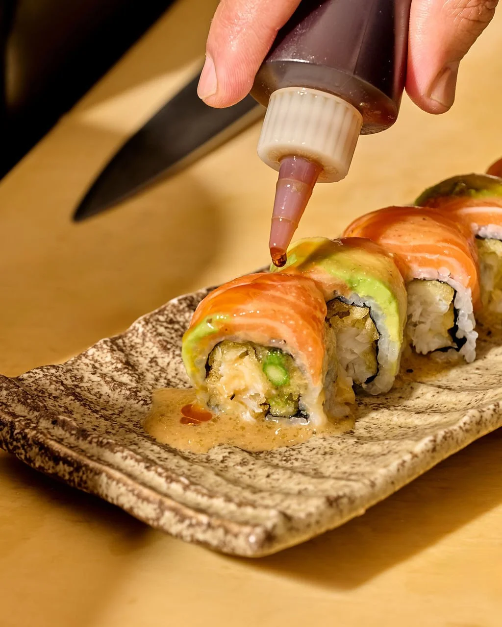 A hand squeezing sauce onto sushi rolls with salmon, avocado, and rice on a decorative plate.
