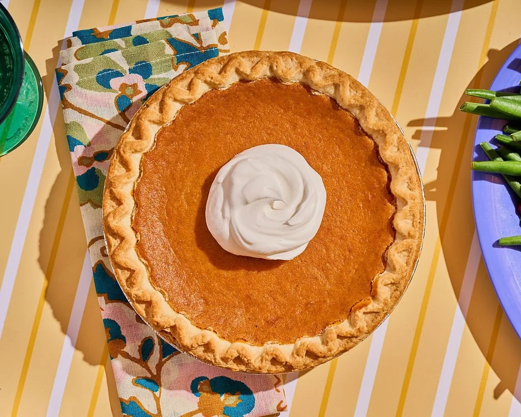 A pumpkin pie with a dollop of whipped cream on top, sitting on a yellow striped table with a colorful cloth and a blue plate of green beans nearby.