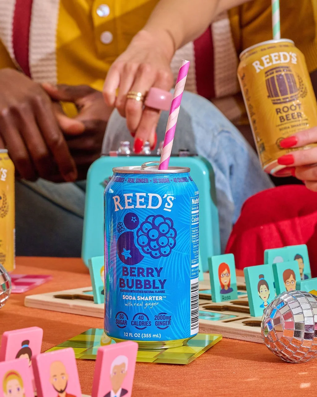 A can of Reed's Berry Bubbly soda with a pink and white striped straw on a table decorated with small illustrated cards and a disco ball themed object, with two people in the background holding cans of Reed's Root Beer.