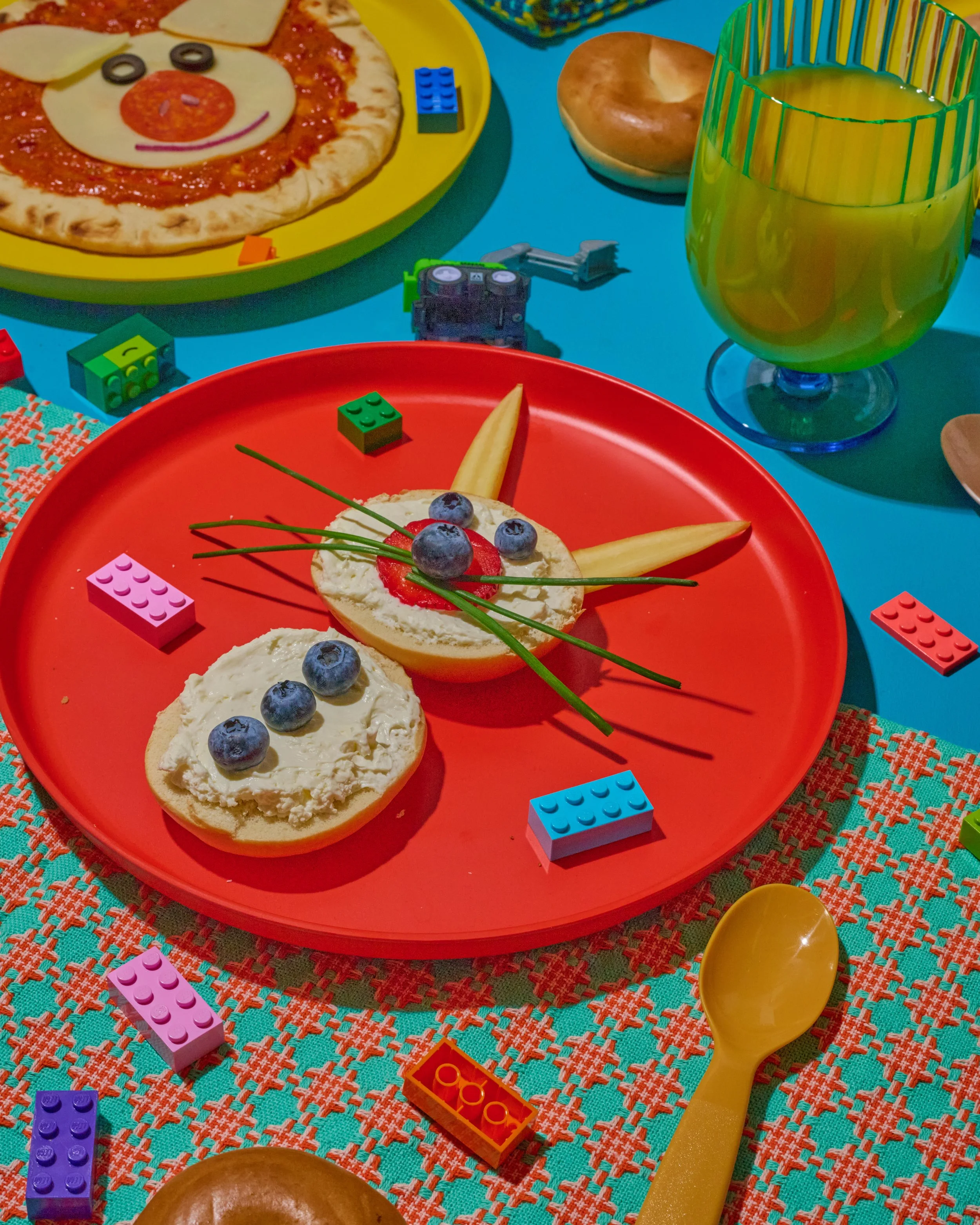 A colorful table setup featuring a Lego-inspired snack and a pizza with face decorations, with a plate of bagels, a glass of yellow-green beverage, and various blocks and toys.