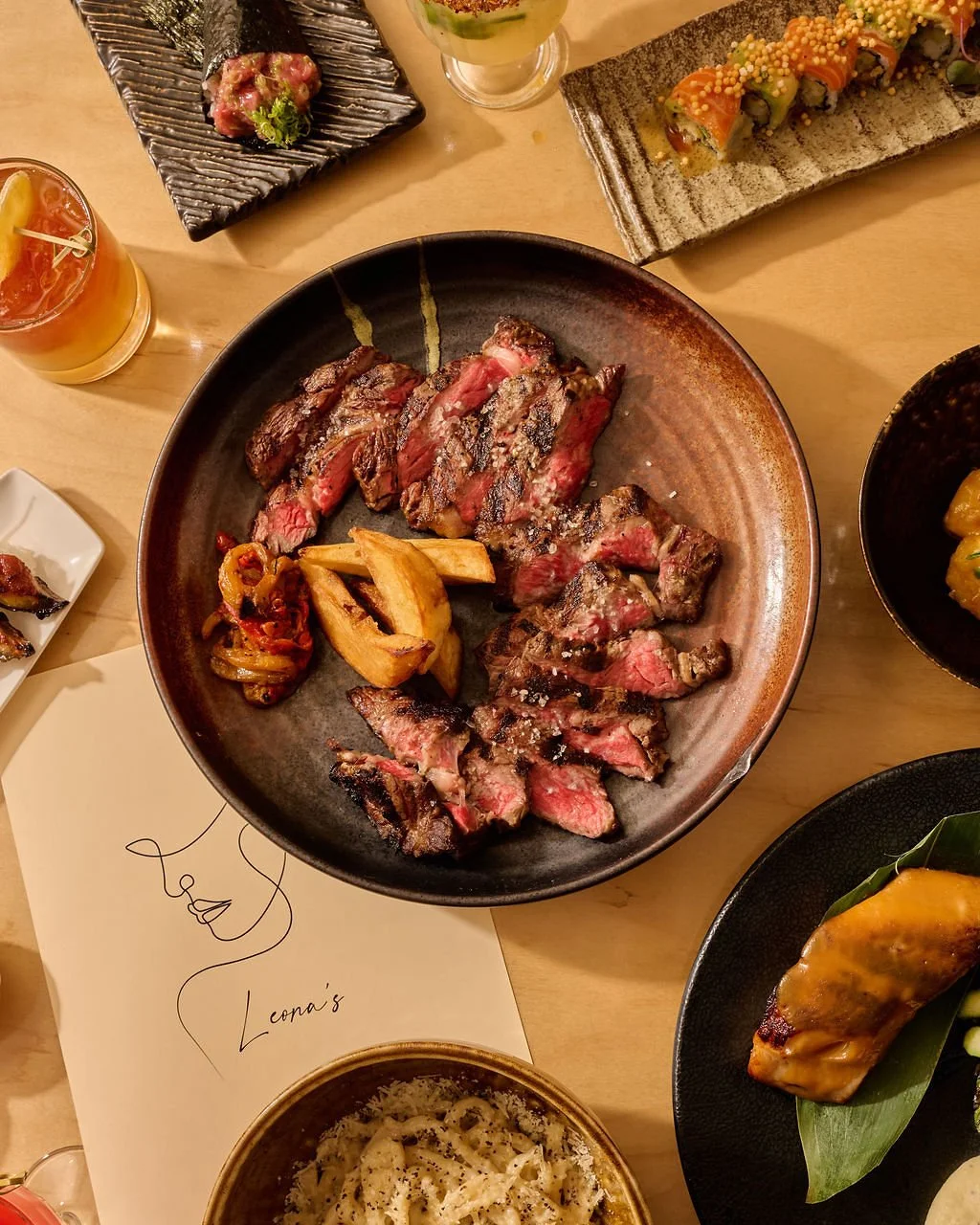 A plate of sliced grilled steak with potato wedges and caramelized onions, surrounded by various Japanese sushi and appetizers on a table.