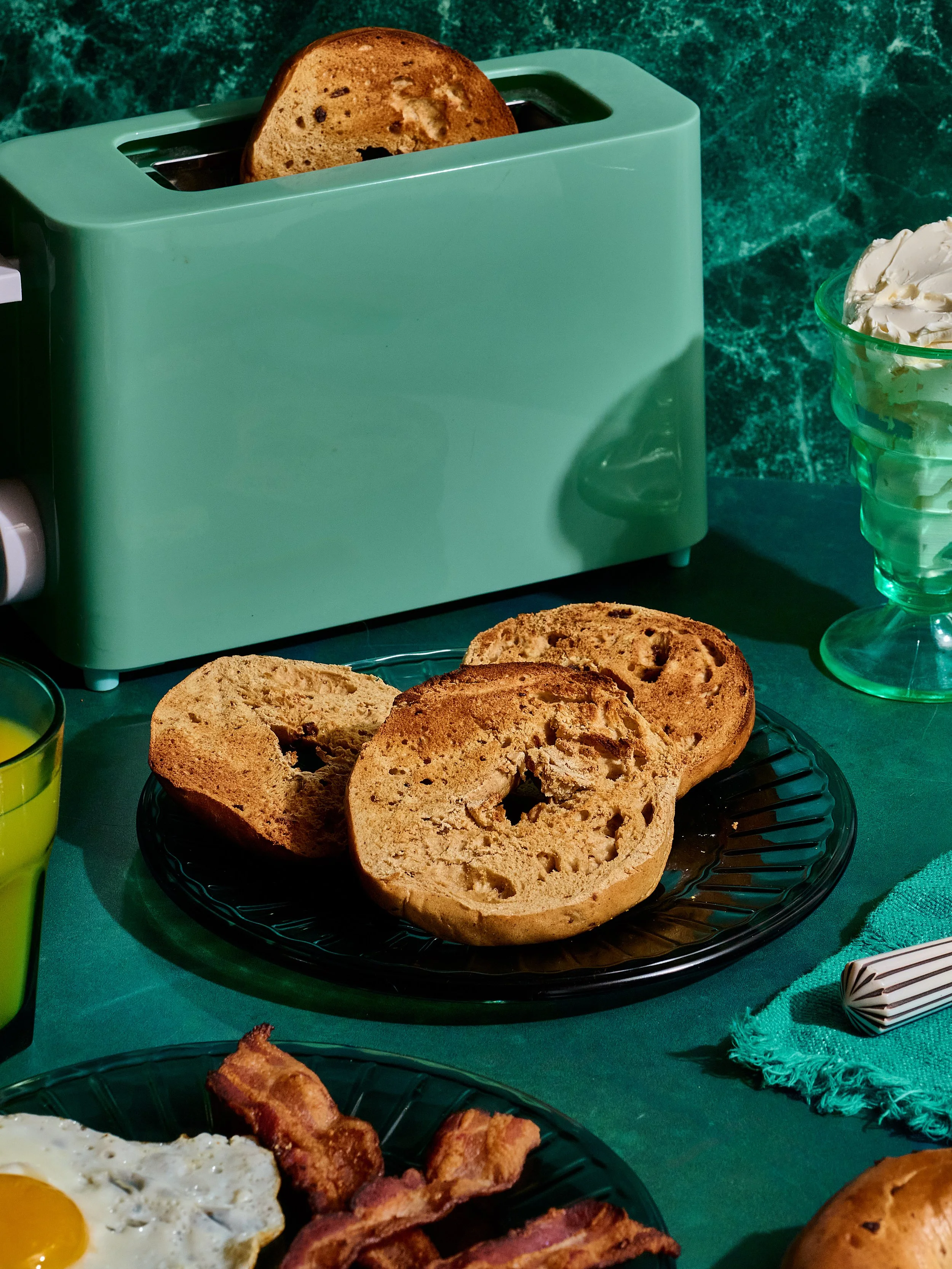 Sliced bagels on a black plate next to bacon and a fried egg at a breakfast table with a teal-tinted drink and a green toaster.