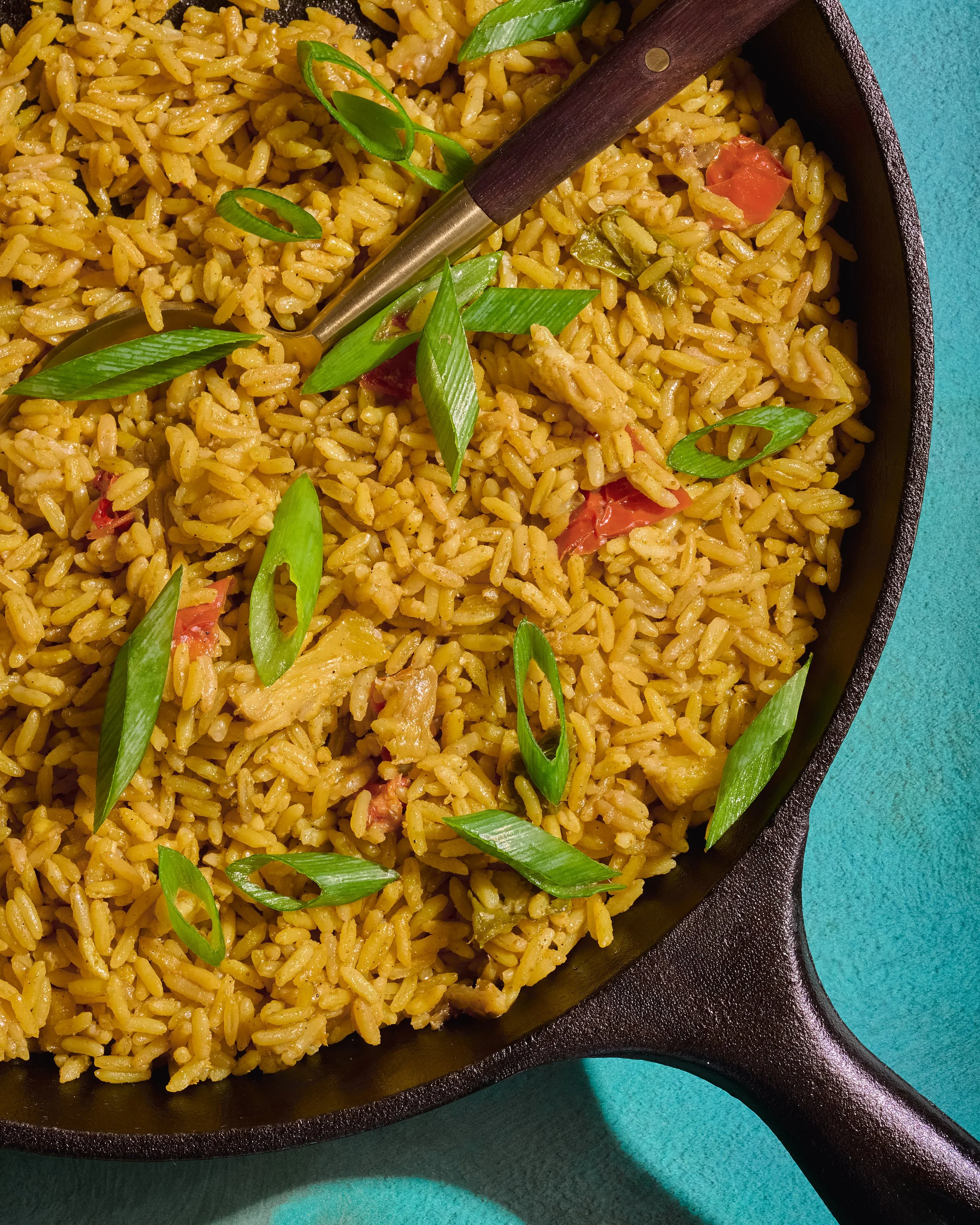 Close-up of a skillet filled with seasoned rice garnished with chopped green onions, with a teal surface in the background.