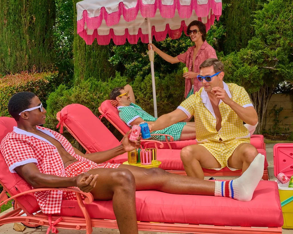Four young people are relaxing on pink lounge chairs in a backyard. They are wearing colorful checkered shirts and sunglasses, enjoying drinks and snacks under a pink and white umbrella, with green bushes and trees in the background.