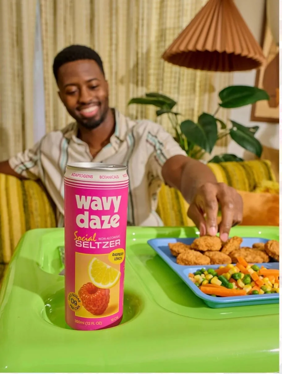 A man sitting at a table with a pink can of Wawy Daze Social Seltzer in front of him, reaching for fried chicken and a plate of mixed vegetables on a blue tray.