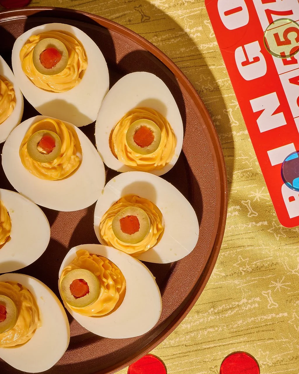 Deviled eggs topped with green olives and red pimiento, on a brown tray in a restaurant setting.