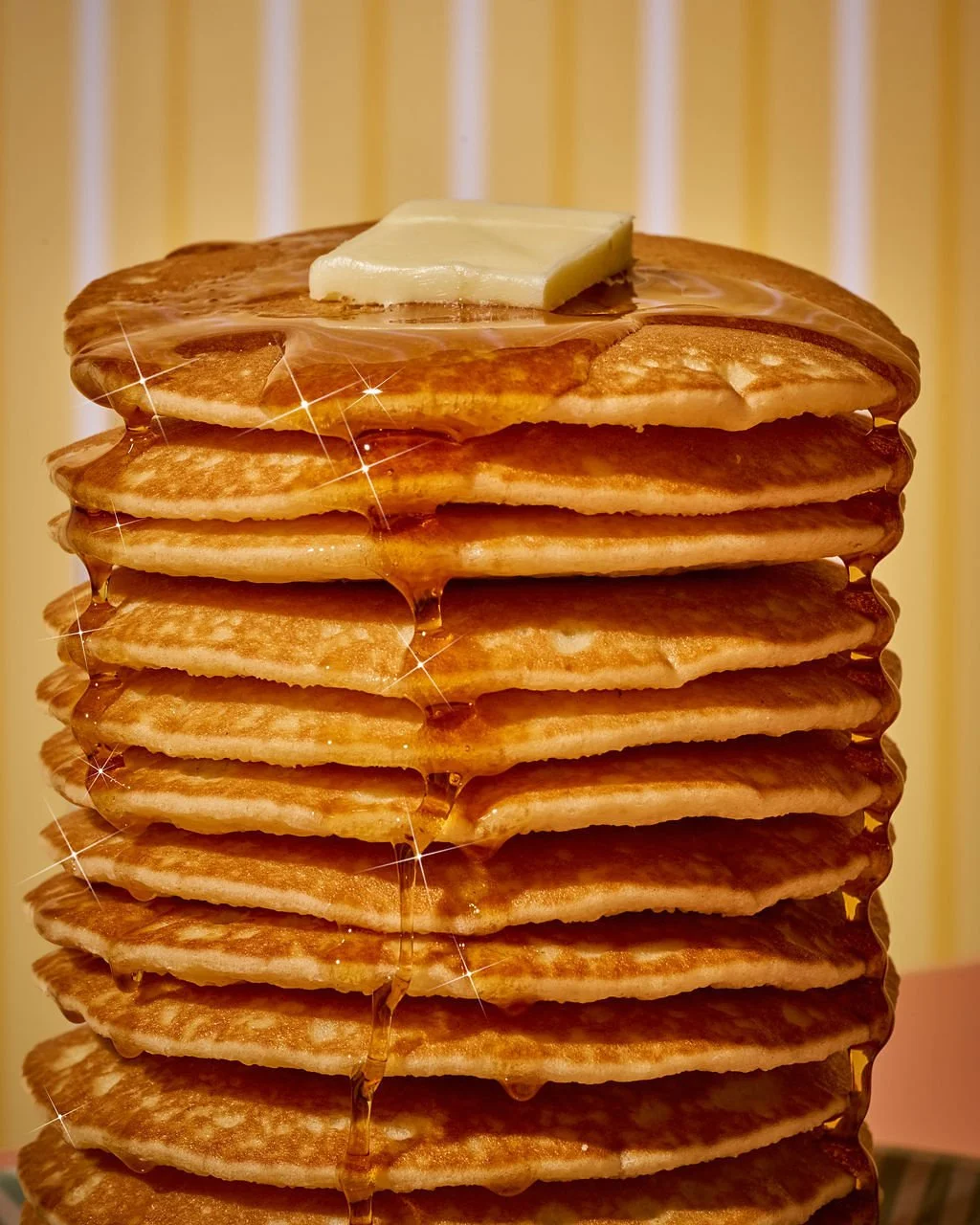 A tall stack of golden brown pancakes with a pat of butter on top, drizzled with syrup, against a patterned yellow background.
