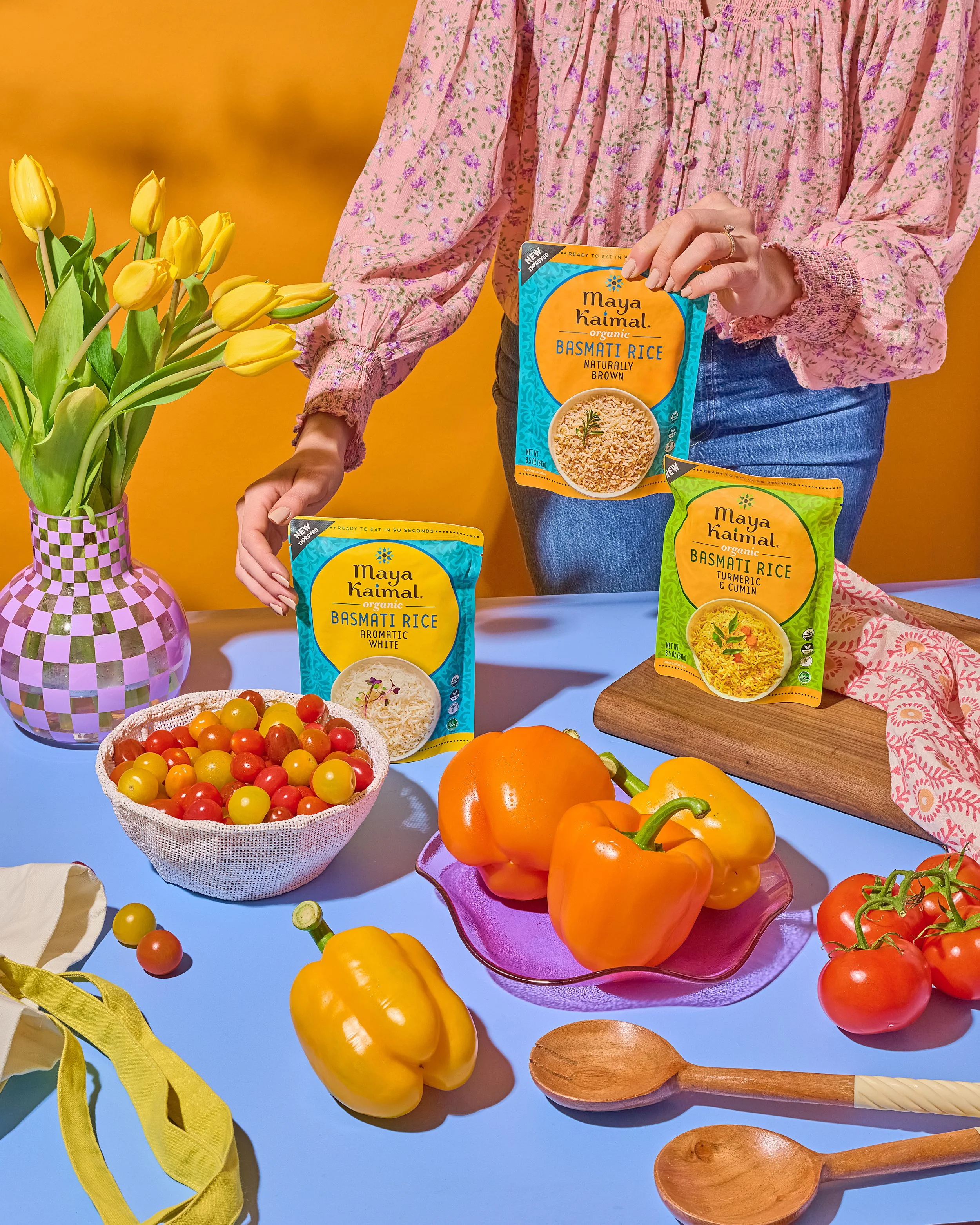 A person holding packages of Maya Kaimal organic basmati rice, surrounded by yellow tulips in a patterned purple and white vase, a bowl of cherry tomatoes, yellow and orange bell peppers, and a wooden cutting board with additional rice packages on a blue table against a yellow background.