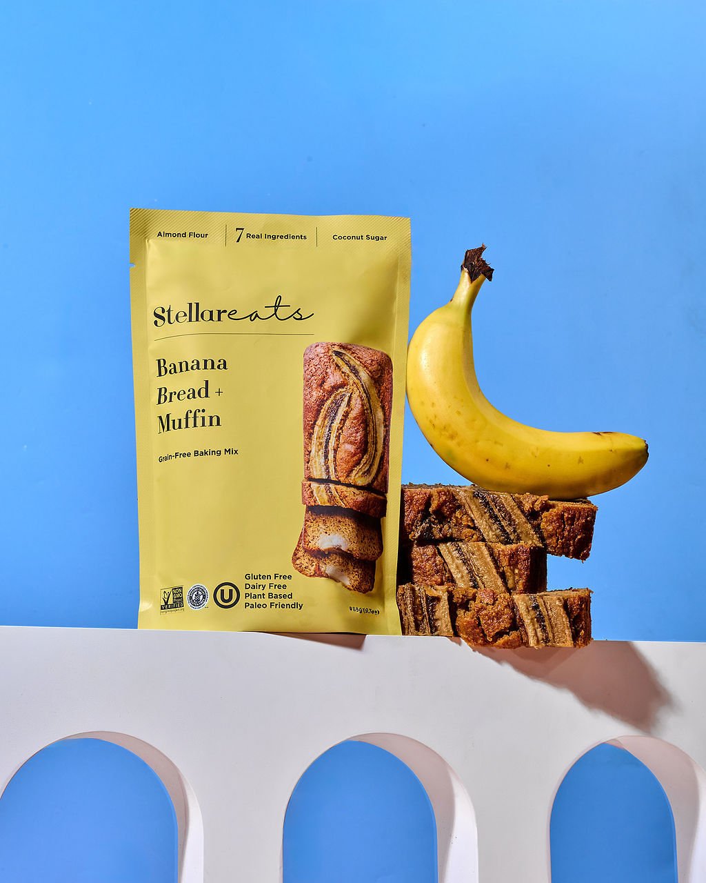 A yellow package of Stellareats Banana Bread Muffin mix, a ripe banana, and several pieces of banana bread with chocolate streaks on top, arranged against a blue background.