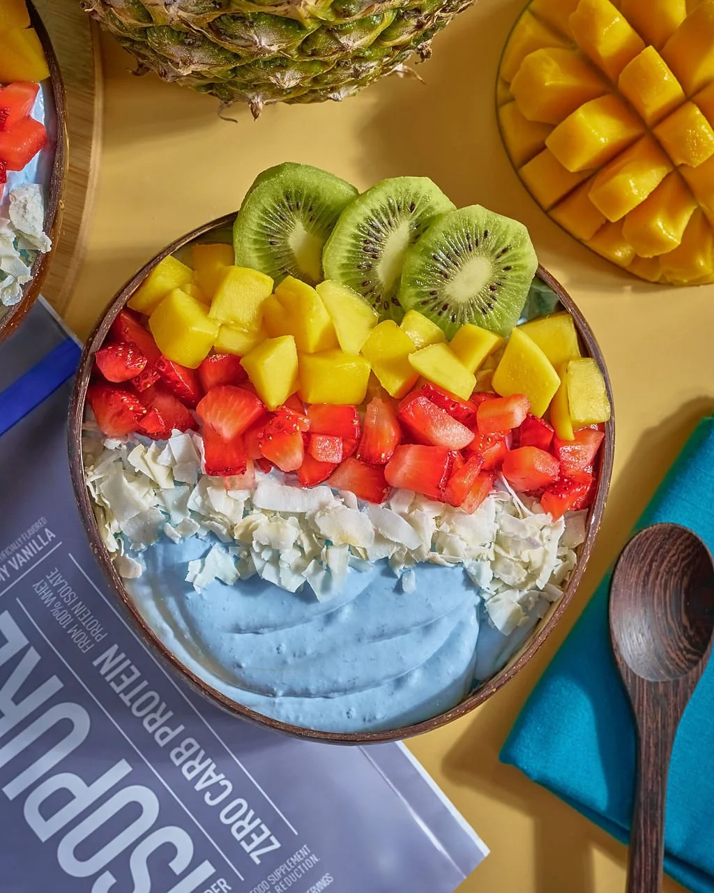 A bowl of blue frosting topped with white chocolate shavings and sliced fresh fruits, including kiwi, mango, strawberries, and pineapple.