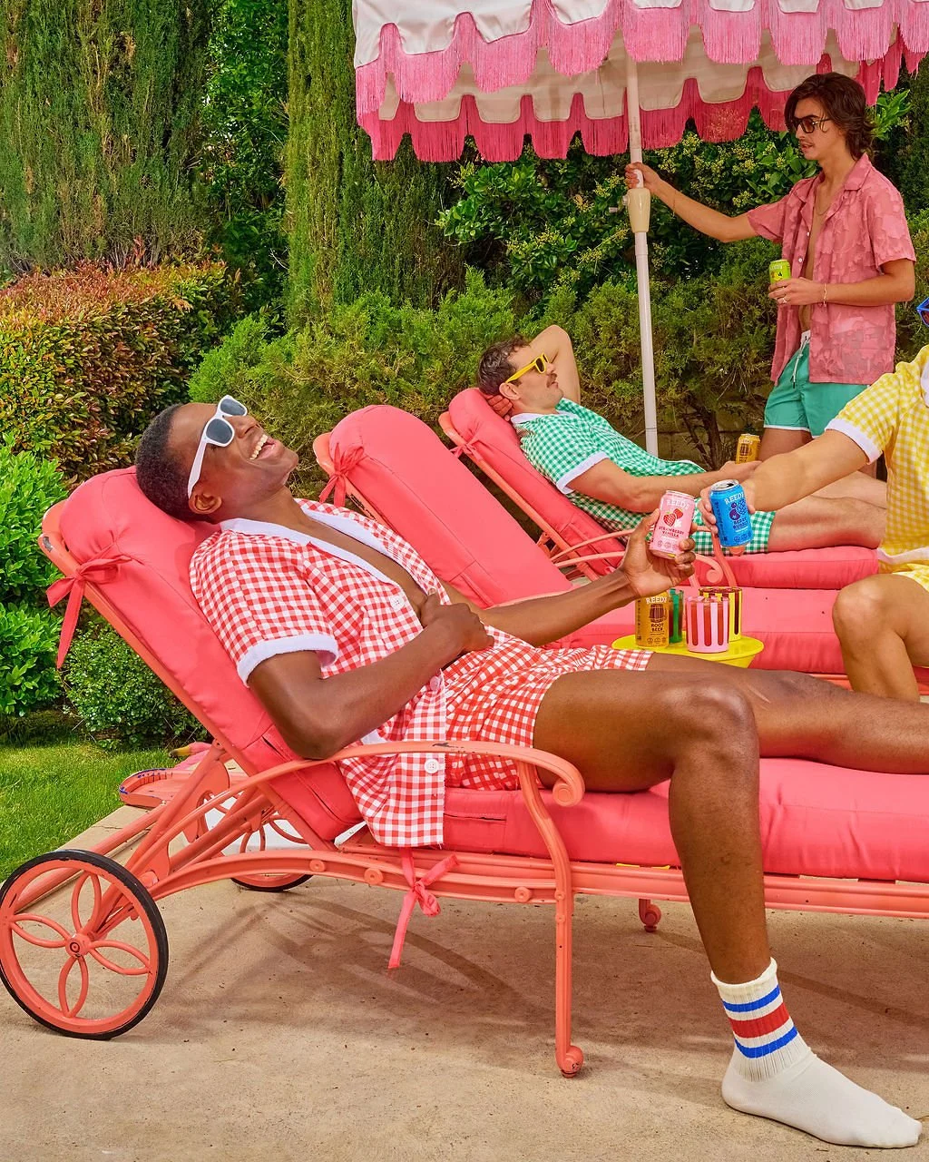 Three people relaxing outdoors on pink lounge chairs under an umbrella, with lush green bushes in the background. One person is smiling and holding a soda can, another is reclining and wearing yellow sunglasses, and a third is standing and holding a drink.