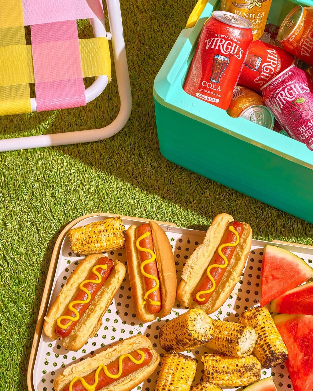 Hot dogs with mustard, grilled corn on the cob, watermelon slices, and canned sodas on a grassy surface with a colorful picnic chair and cooler.