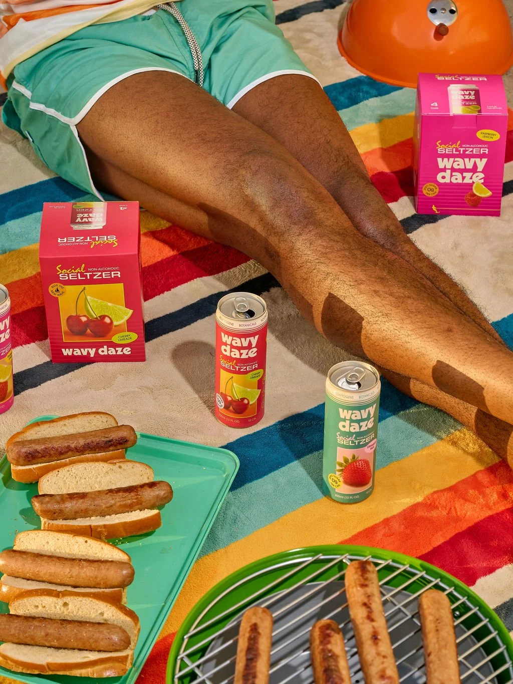 Person lying on a colorful striped blanket with legs extended, surrounded by boxes of Wavy Daze seltzer, a green plate with hot dogs, and a green skillet with cooked sausages, with a small orange toy and a few soda cans nearby.