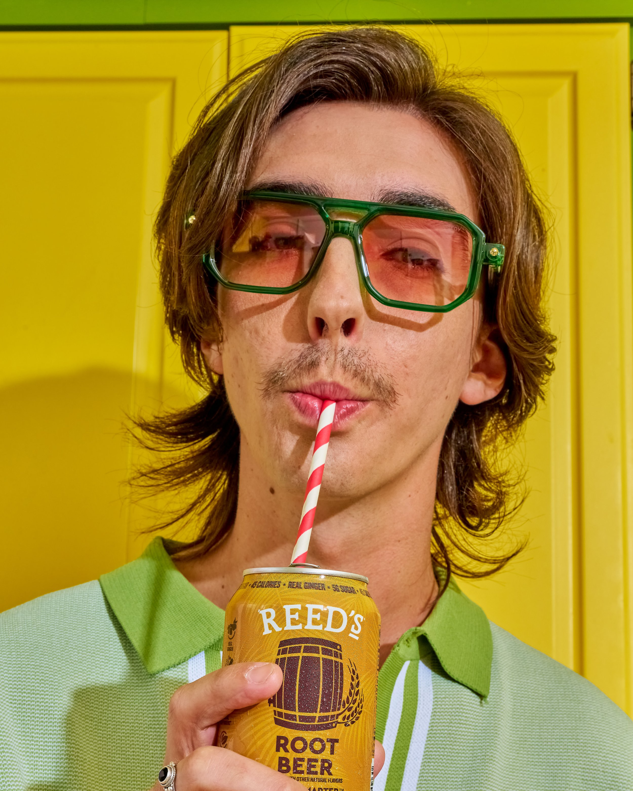 A man with medium-length brown hair, wearing oversized green sunglasses, a green shirt, and a green vest, is sipping from a can of Reed's Root Beer through a red and white striped straw, standing in front of a bright yellow background.