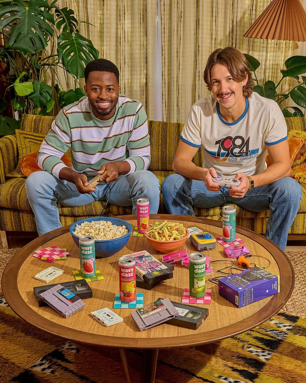 Two men sitting on a retro yellow-green patterned couch playing video games, with snacks and gaming items on the coffee table in front of them, in a cozy living room with plants and curtains.