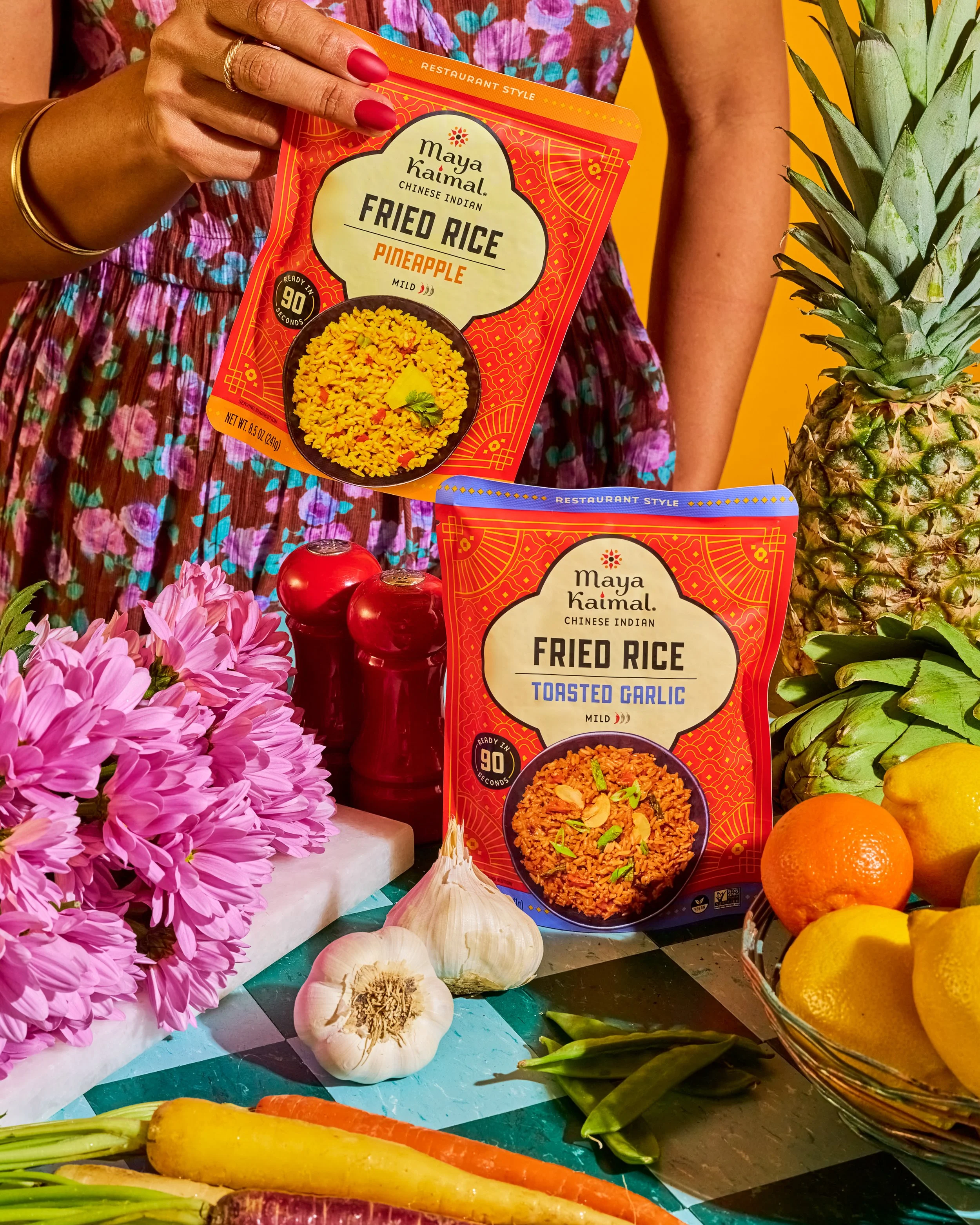 Person holding a box of Maya Kaimal Thai fried rice, with fresh vegetables and flowers around, including garlic, carrots, an orange, and a pineapple, against a colorful background.