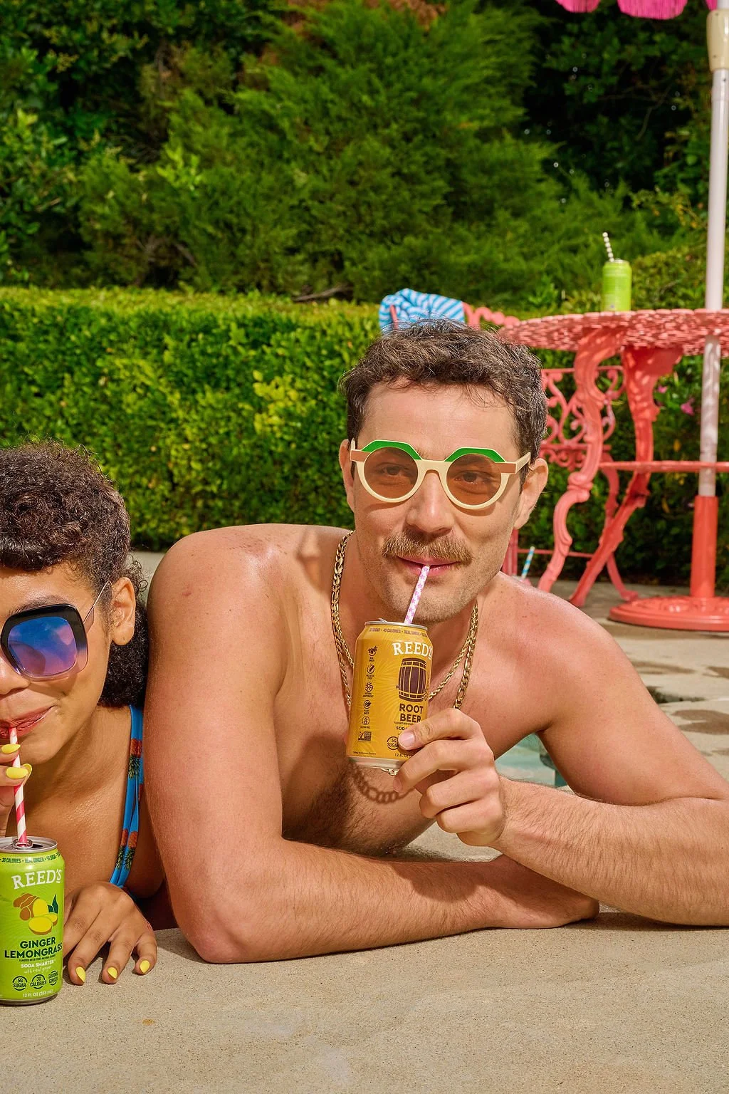 A shirtless man with sunglasses and a mustache relaxing at a poolside, holding a can of Reed's Root Beer with a straw, next to a girl with sunglasses drinking a ginger lemonade soda.