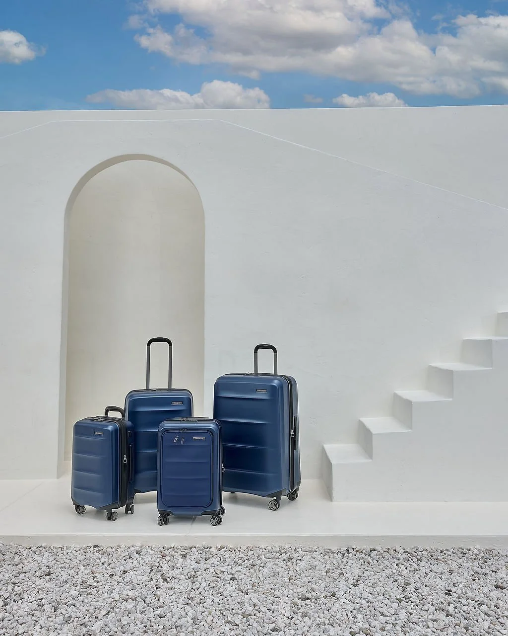Four blue suitcases of different sizes on a white surface against a white wall with stairs and an arched doorway, under a partly cloudy sky.