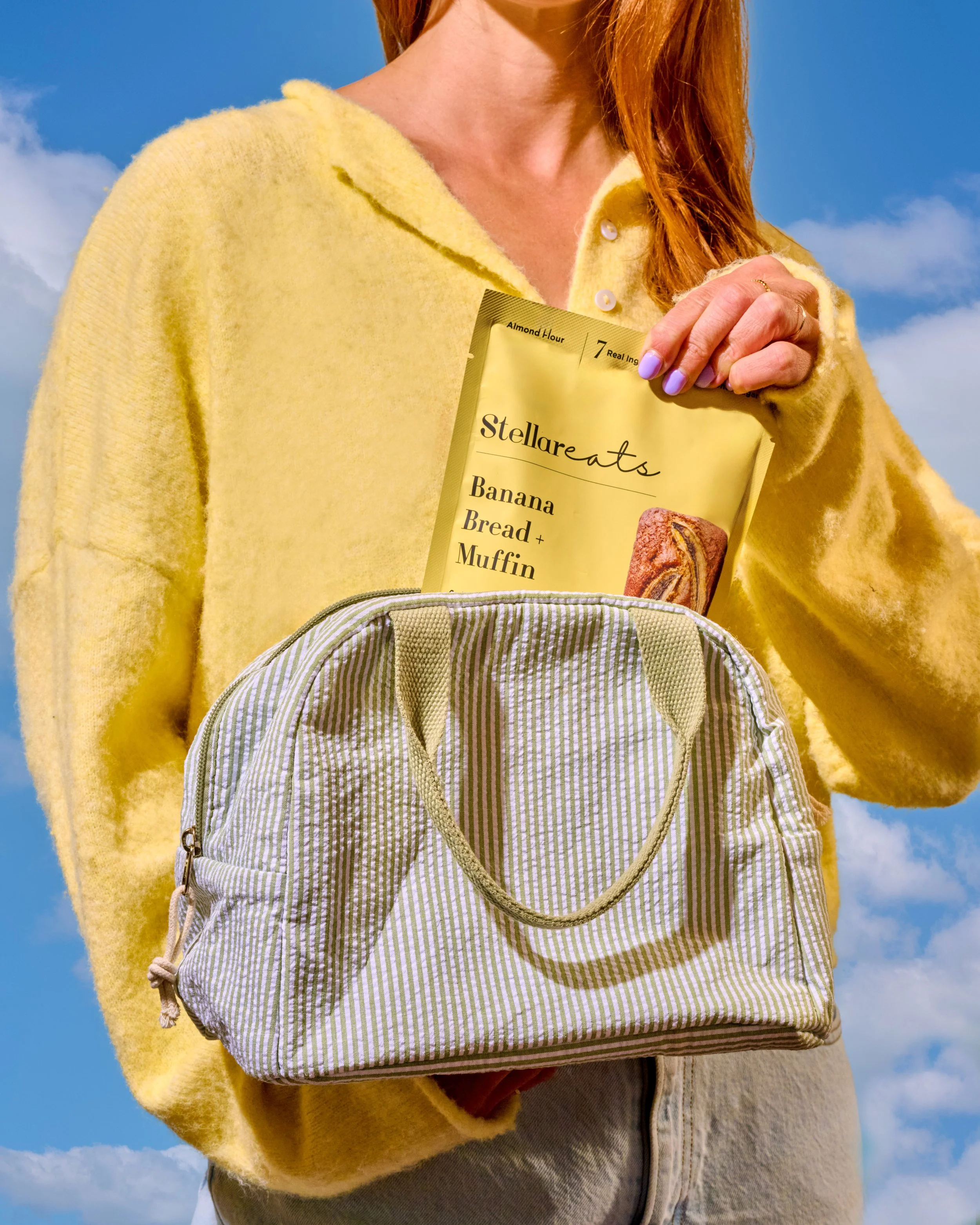 A person wearing a yellow cardigan holding a yellow box of Stellaeats banana bread and muffin mix, with a blue sky and clouds in the background.