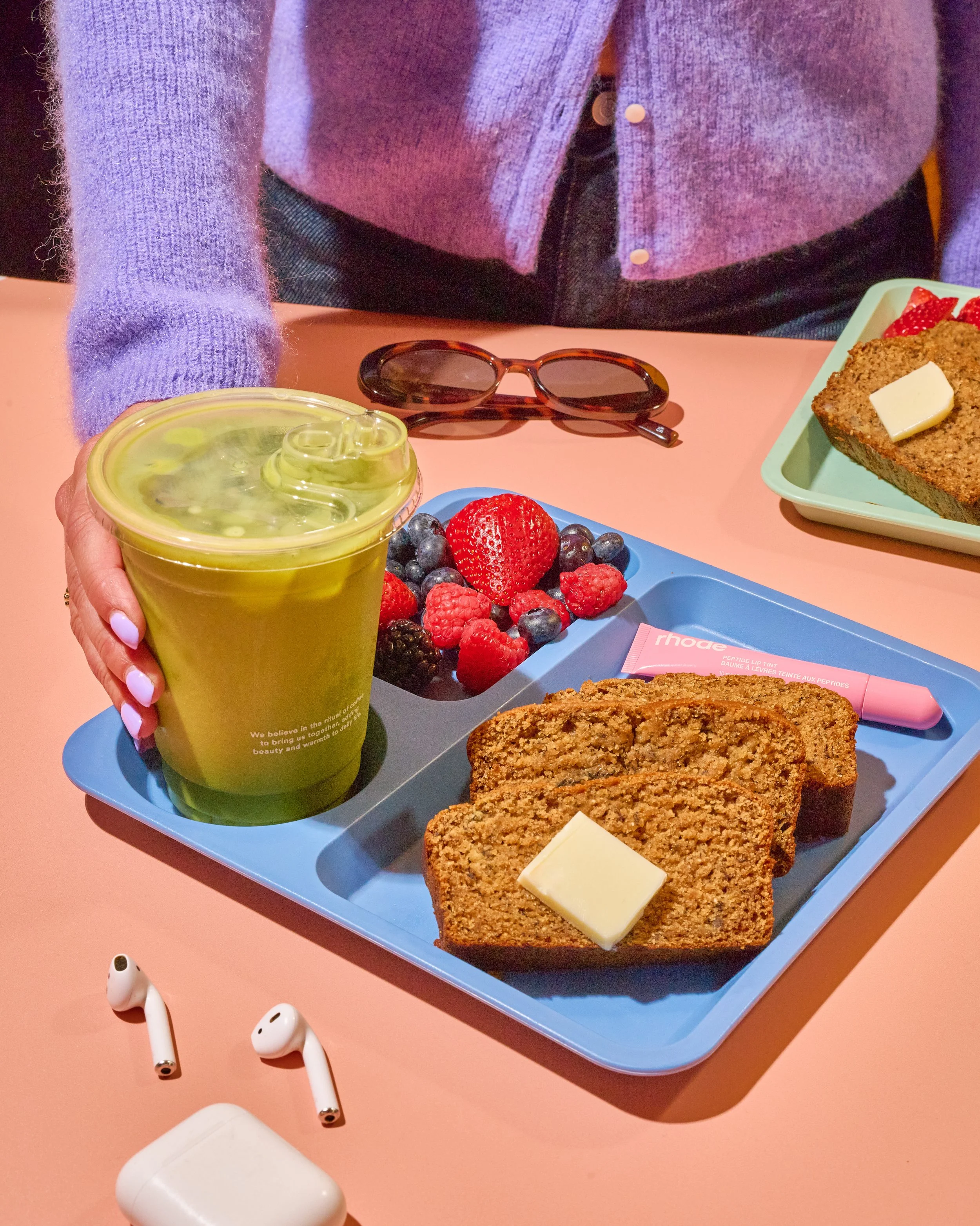 A person holding a green drink with ice, sitting at a pink table with a blue tray of food including strawberries, blueberries, raspberries, two slices of banana bread with butter, pink lip balm, a pair of glasses, and a second plate of banana bread with butter in the background.