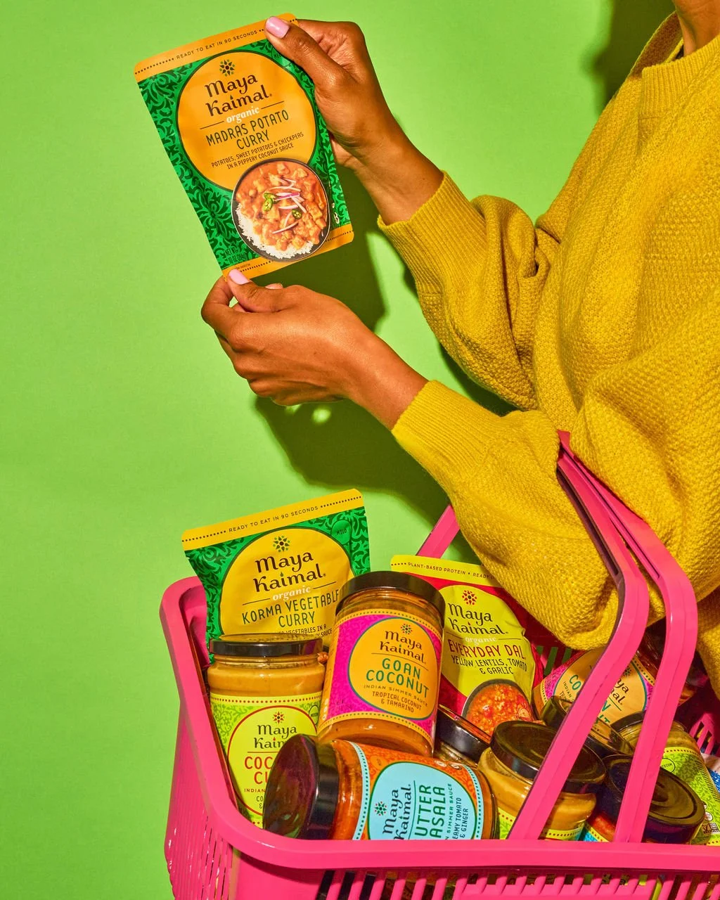 A person in a yellow sweater holding a Maya Kaimal organic curry package against a green background, with a pink shopping basket filled with various Maya Kaimal products on a green table.