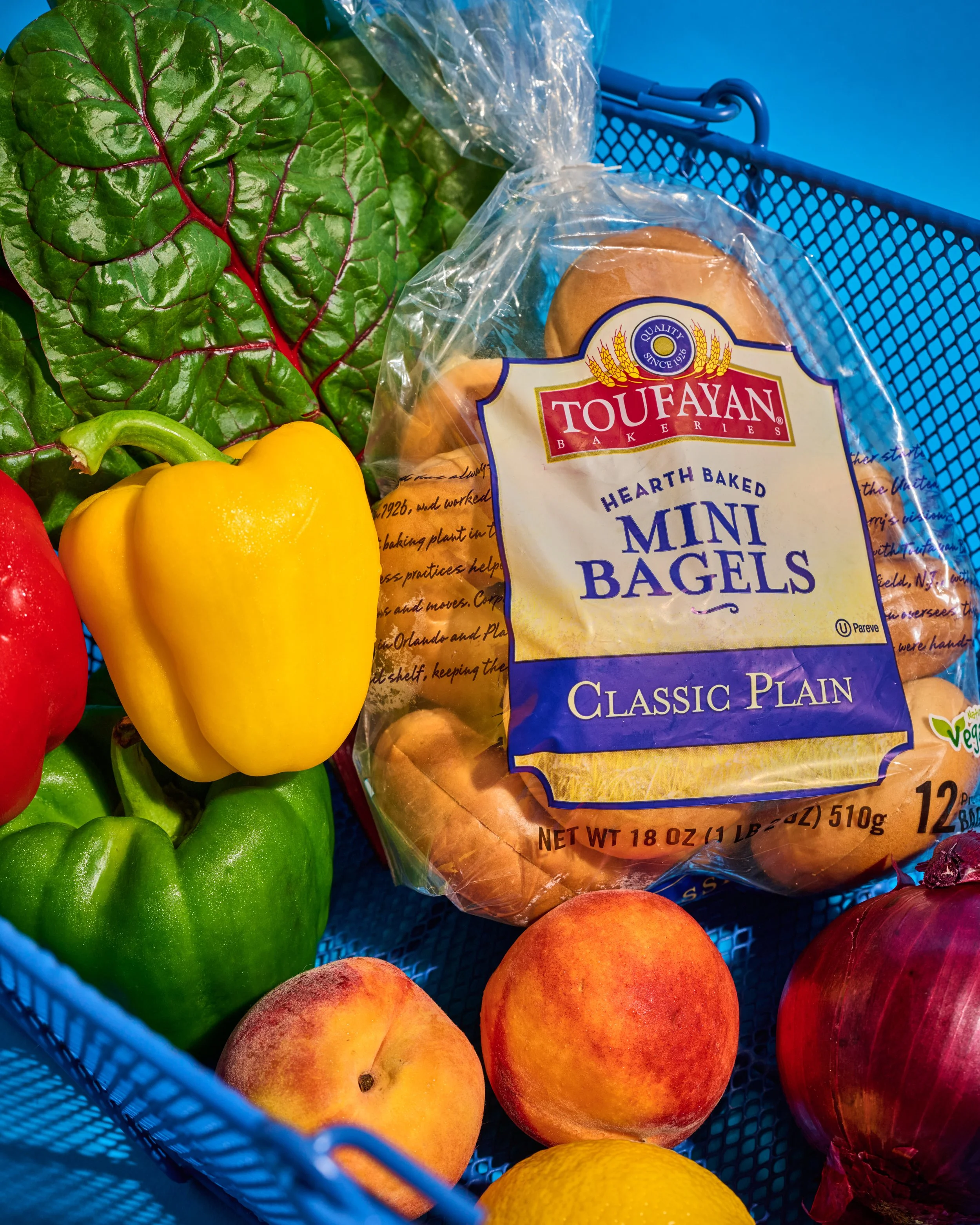 A shopping basket containing freshly picked fruits and vegetables, including green and yellow bell peppers, peaches, and leafy greens, with a pack of mini bagels in the background.